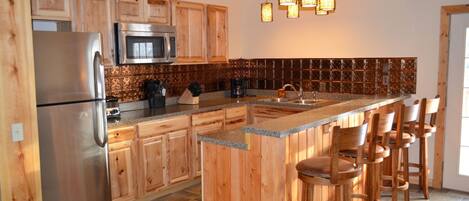 A kitchen with wooden cabinets, a stainless steel refrigerator, a microwave, a coffee maker, and a bar area with stools.