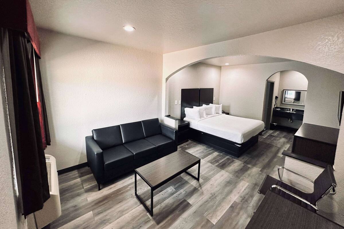 A modern hotel room with a black sofa, a bed with white linens, a small table, and a bathroom with a mirror and sink.