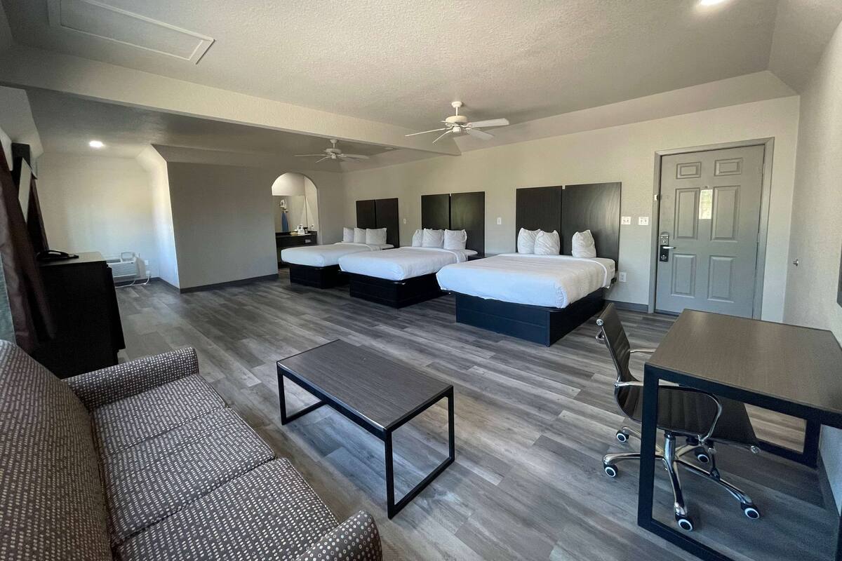 A hotel room with two beds, a desk, a chair, a sofa, and a ceiling fan.