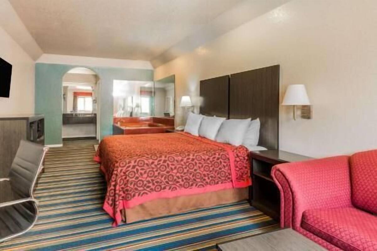 A hotel room with a bed, a TV, a desk, a chair, and a bathroom visible in the background.