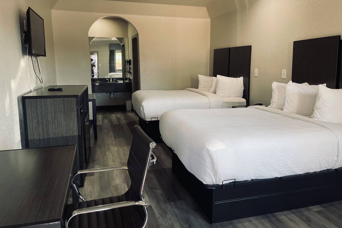 A hotel room with two beds, a desk, a chair, a TV, and a bathroom visible through an archway.