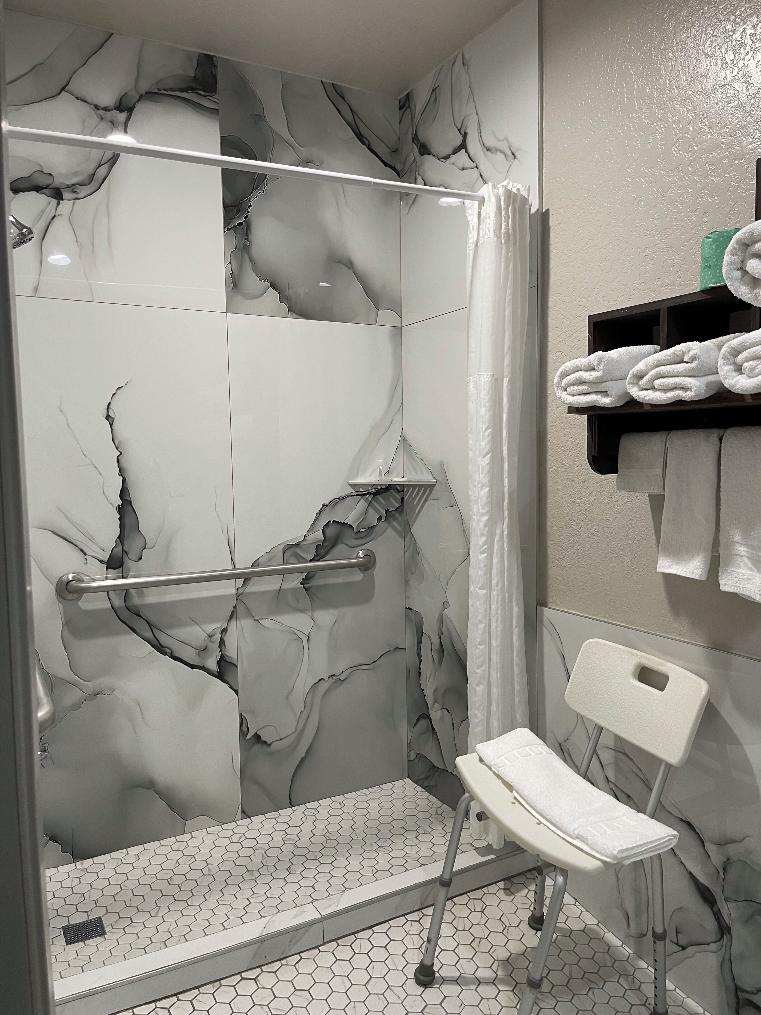 A bathroom with a shower area featuring a marble-like wall design, a white chair, and a shelf with towels.