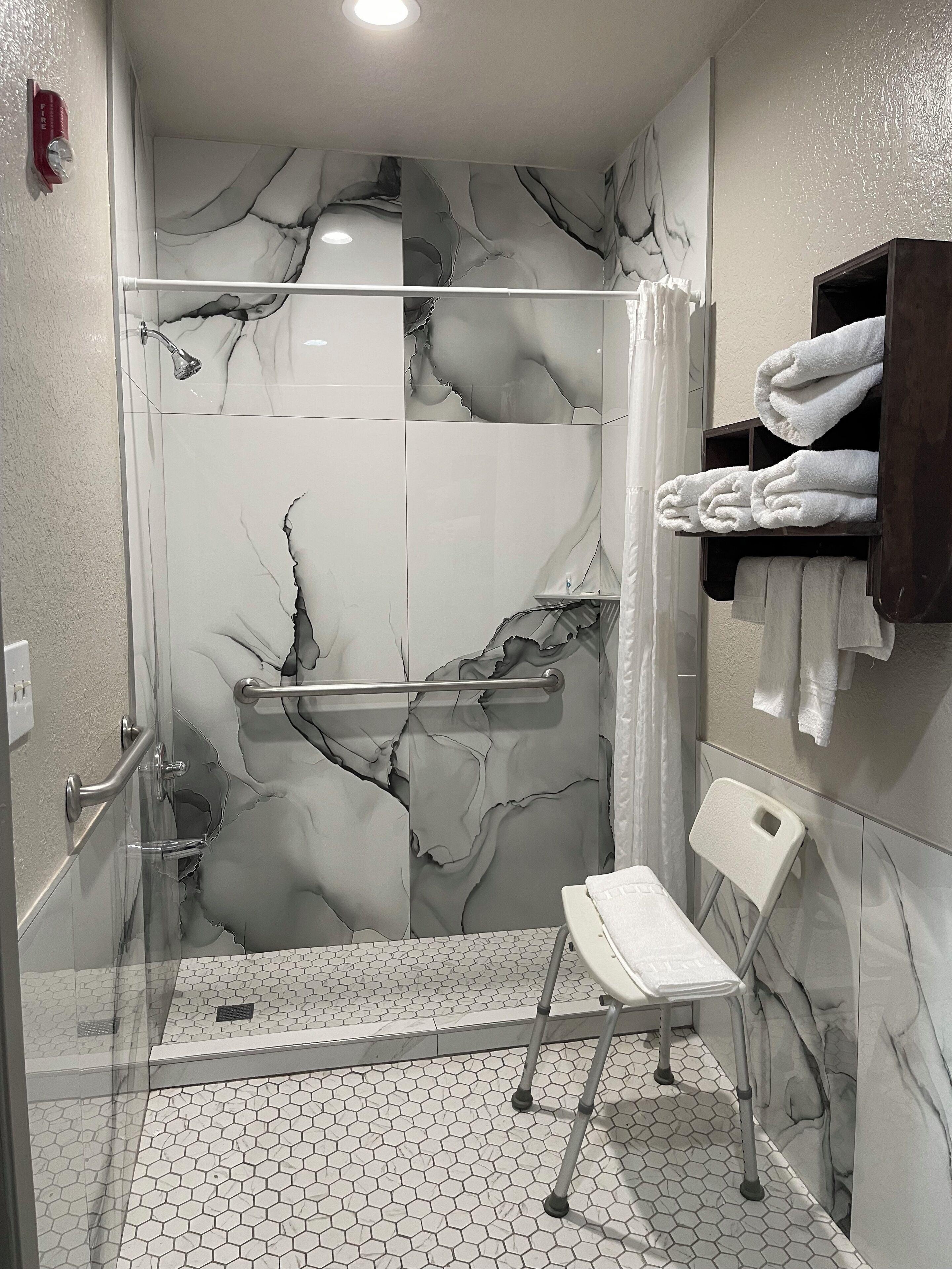 A modern bathroom with a glass-enclosed shower, marble wall tiles, a white hexagonal floor pattern, a white chair, and a towel rack with folded towels.