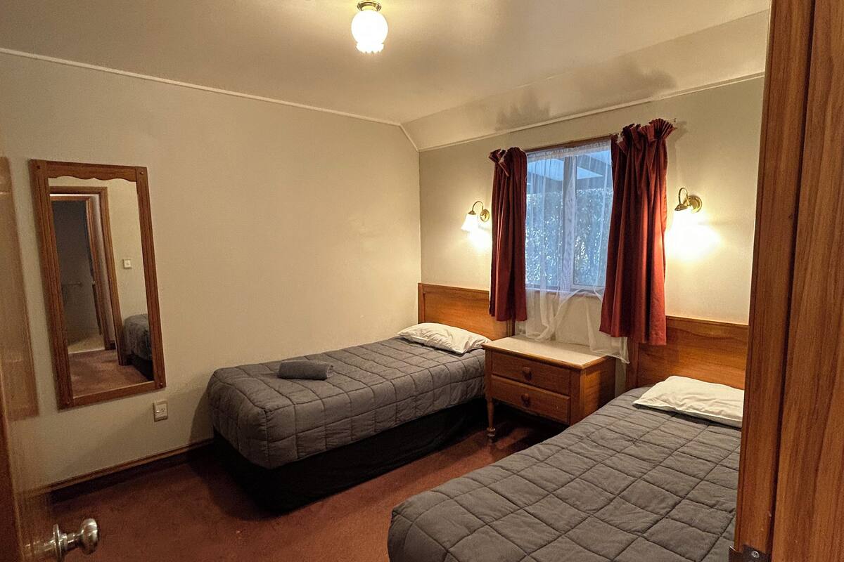 Habitación de hotel con dos camas individuales, una mesita de noche de madera, un espejo y una ventana con cortinas.
