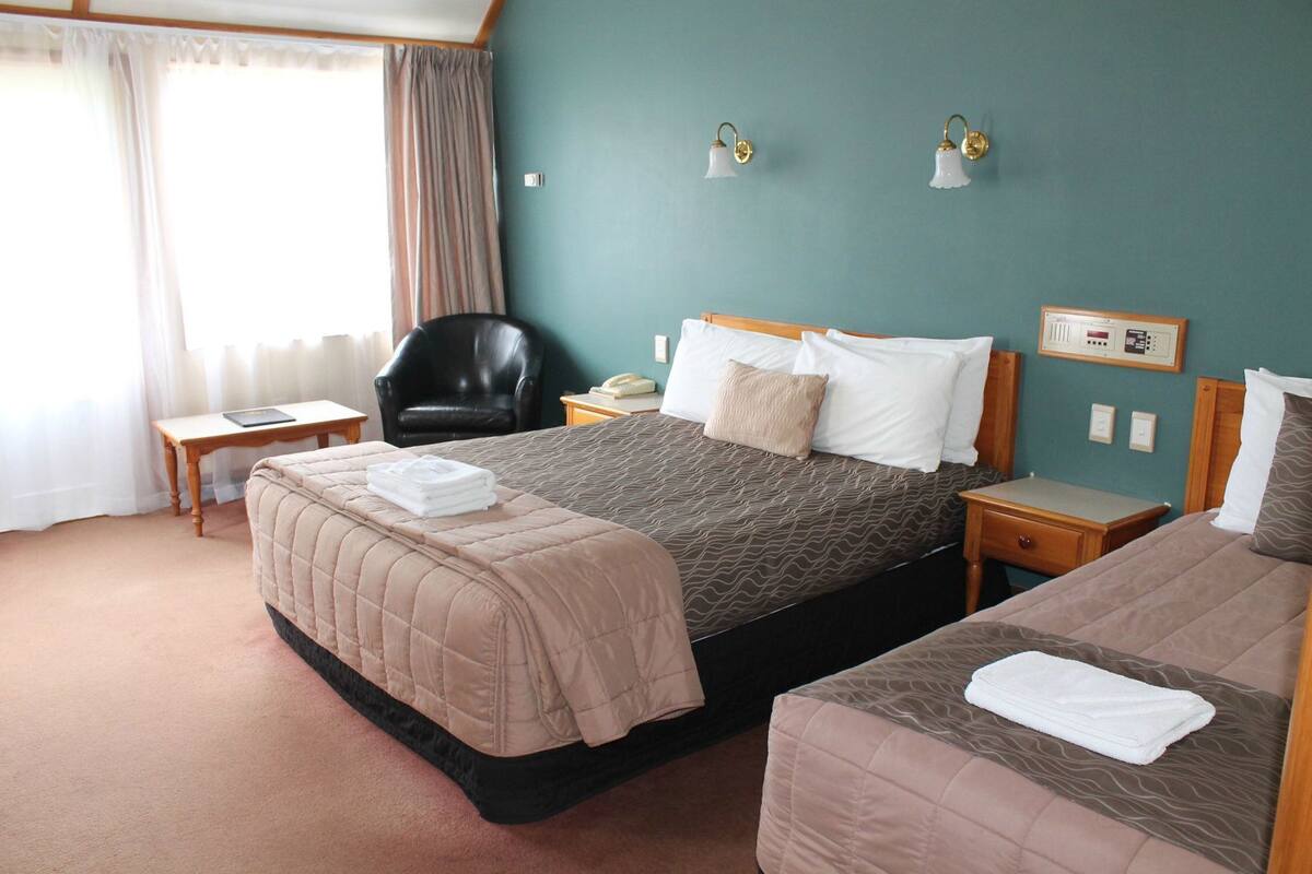 Habitación de hotel con una cama grande, una cama más pequeña, un escritorio, una silla y una mesita de noche con lámpara.