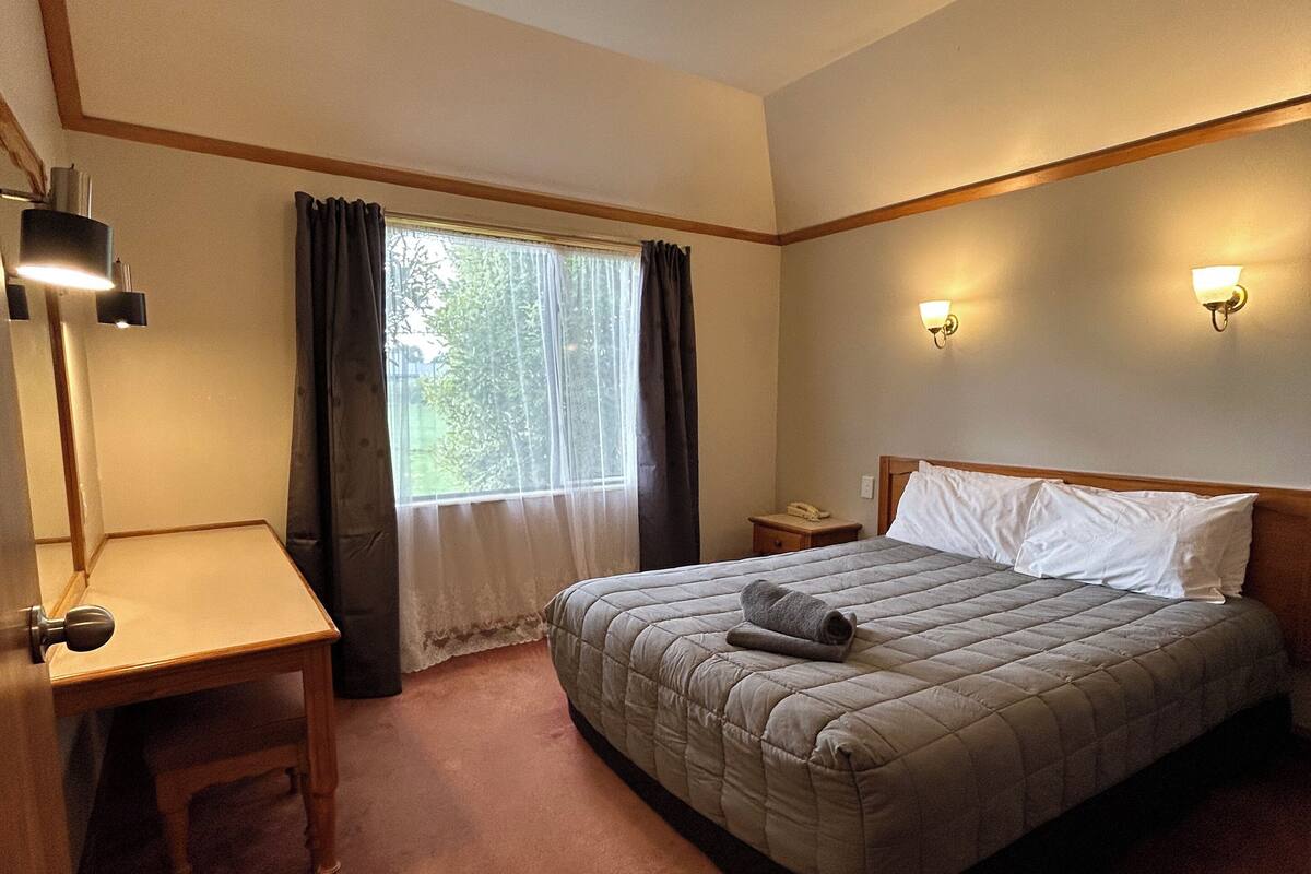Un dormitorio con cama, mesita de noche, lámpara, ventana con cortinas e iluminación en la pared.