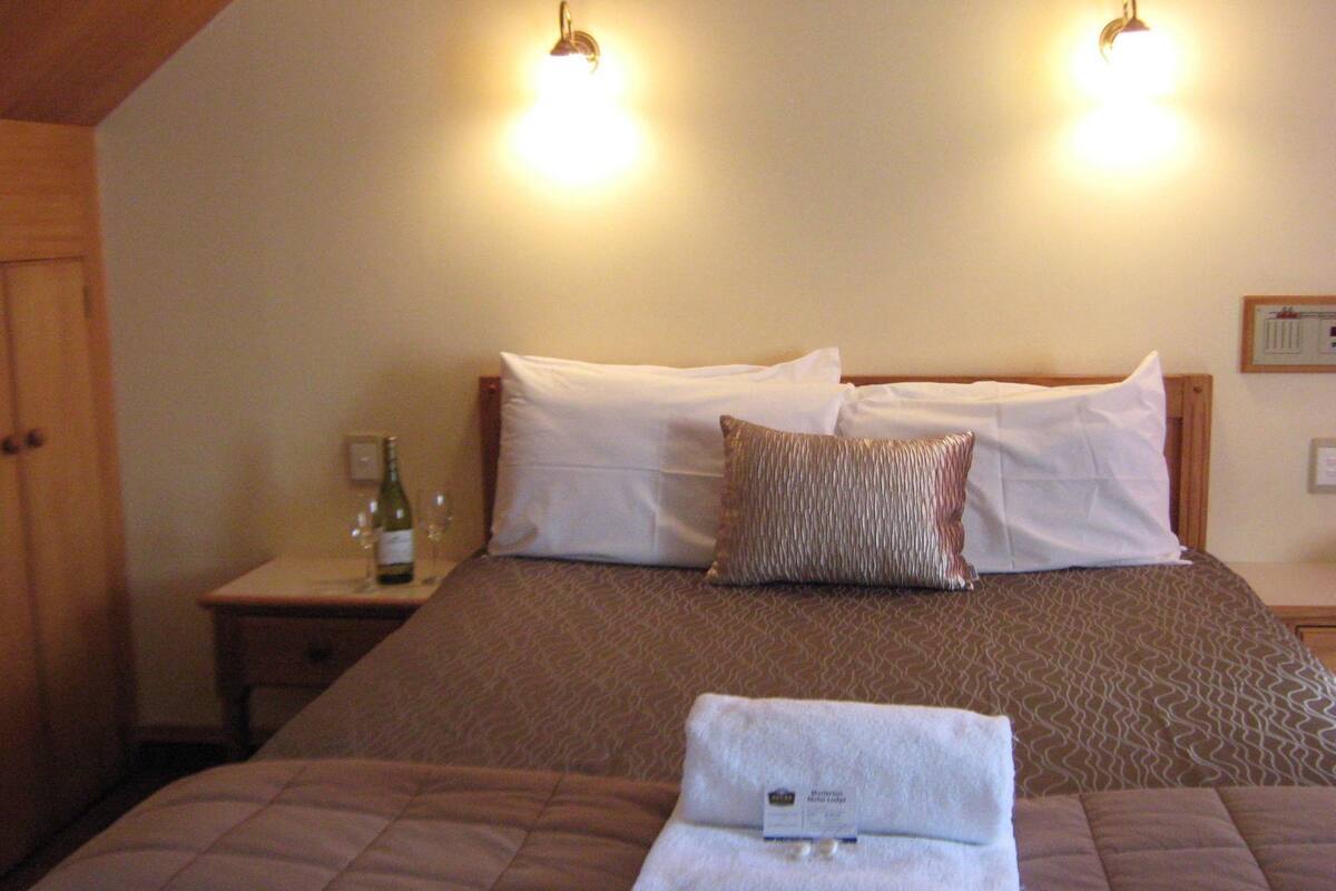 Una habitación de hotel con una cama, una mesita de noche con una botella y vasos, y una toalla doblada sobre la cama.