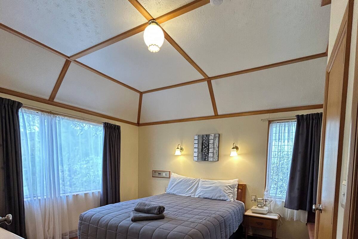 Un dormitorio con cama, mesitas de noche, lámpara de techo y ventana con cortinas.
