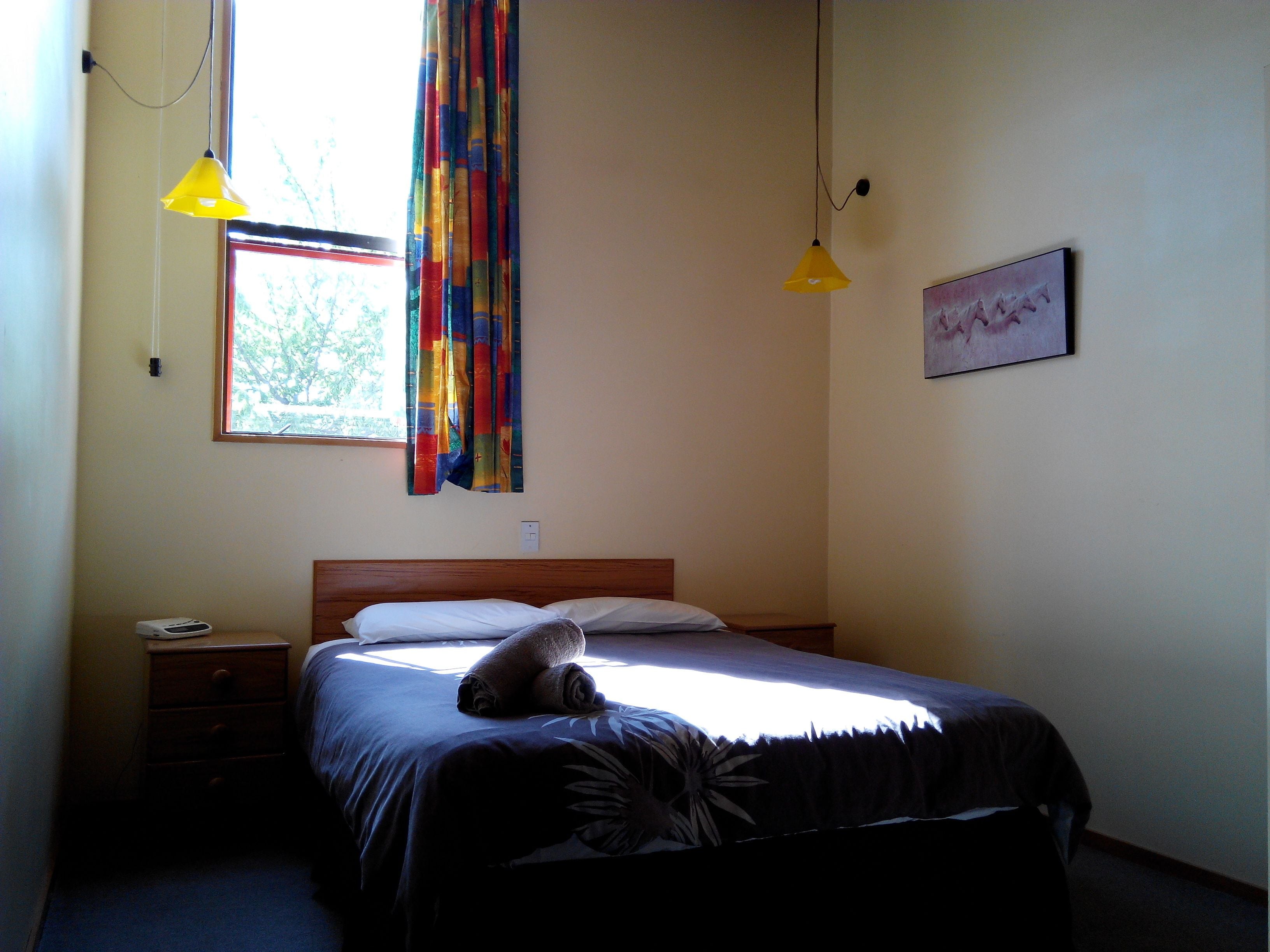 Un dormitorio con una cama, dos mesitas de noche, una ventana con cortinas y dos lámparas colgantes.