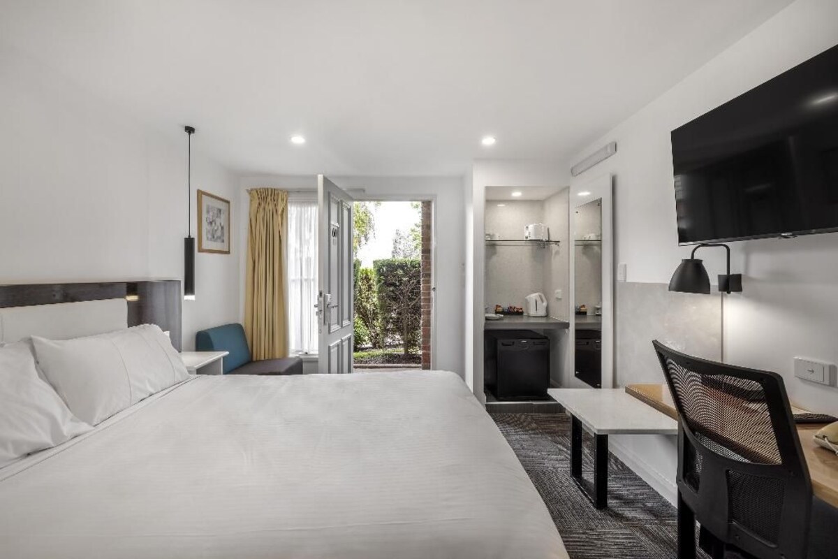 A hotel room with a large bed, a desk, a chair, a TV, and a kitchenette.