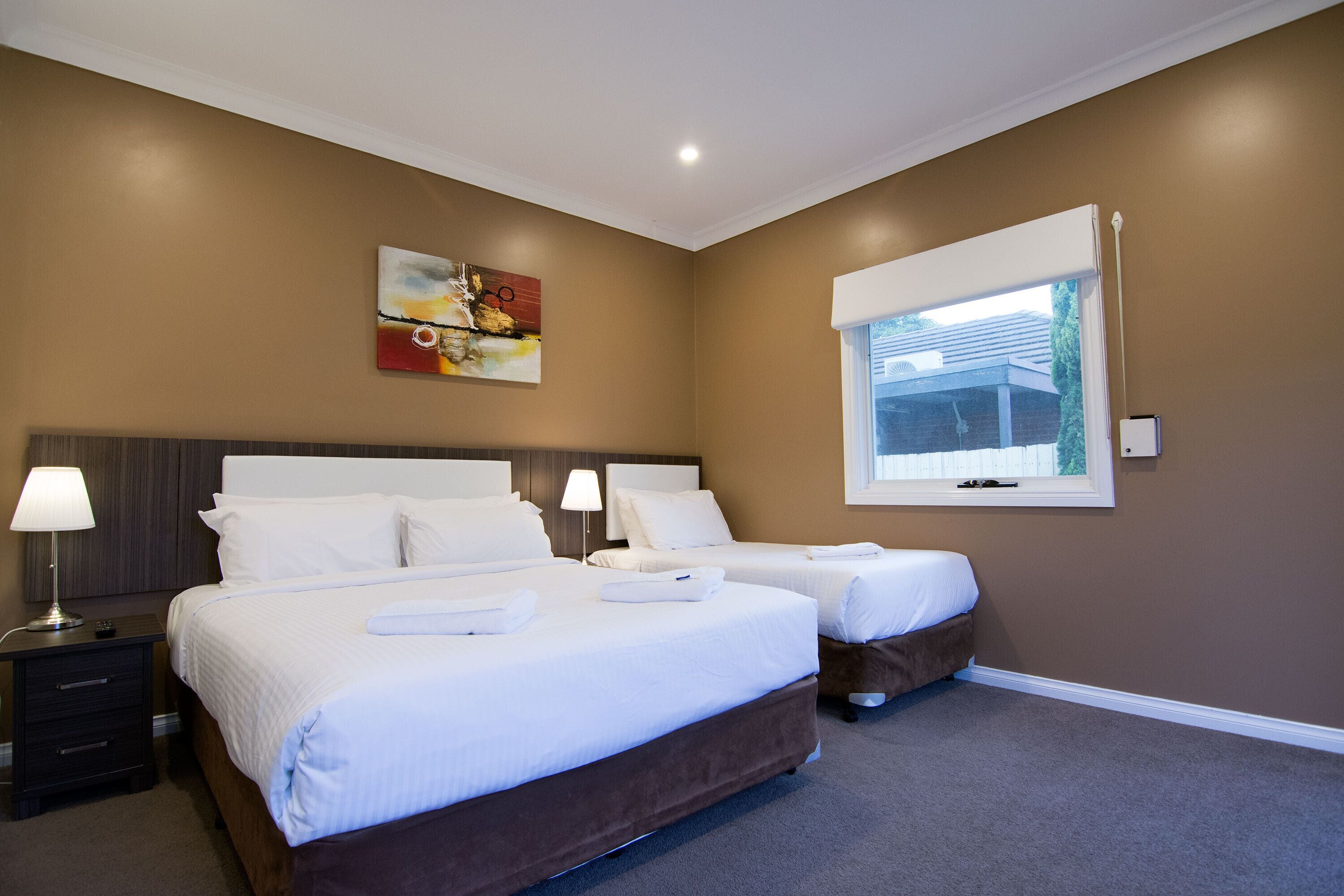A hotel room with two beds, a nightstand, a lamp, a painting on the wall, and a window with blinds.