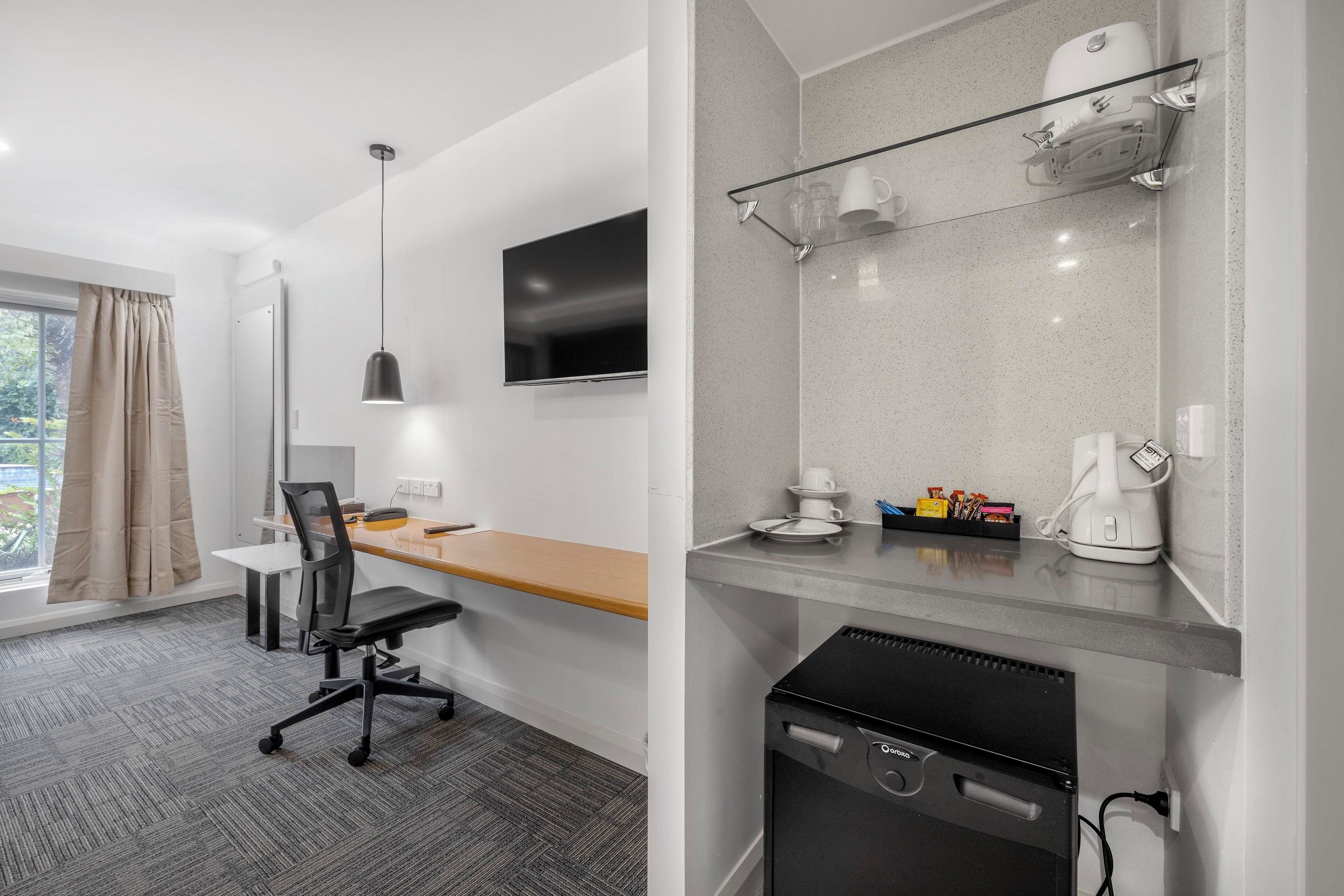 A modern hotel room with a wooden desk, a black office chair, a flat-screen TV, a coffee machine, and a dishwasher.