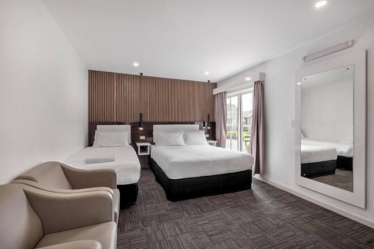Habitación de hotel con dos camas, un sofá, un espejo grande y vistas al exterior.