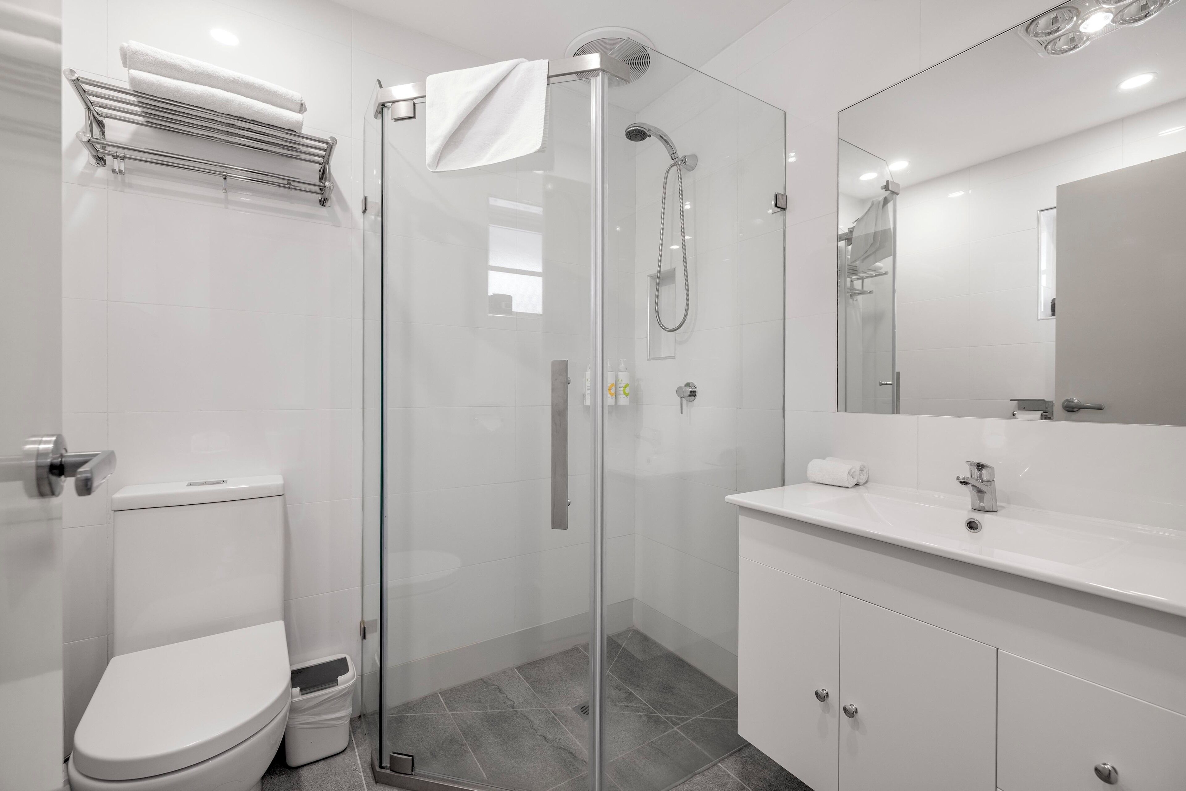 Un baño moderno con ducha envuelta en vidrio, inodoro blanco, tocador con espejo y perchero para toallas.
