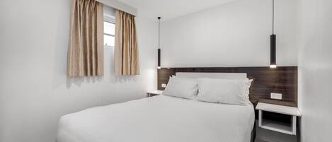 Quarto de hotel moderno com uma cama grande, mesa de cabeceira, cortinas e uma luminária pendente.