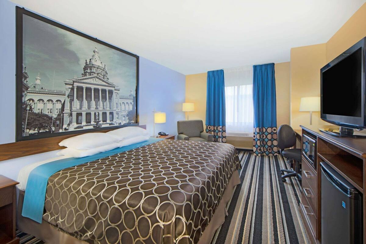 Een hotelkamer met een groot bed, een bureau met een computer, een televisietoestel en een schilderij van een gebouw aan de muur.