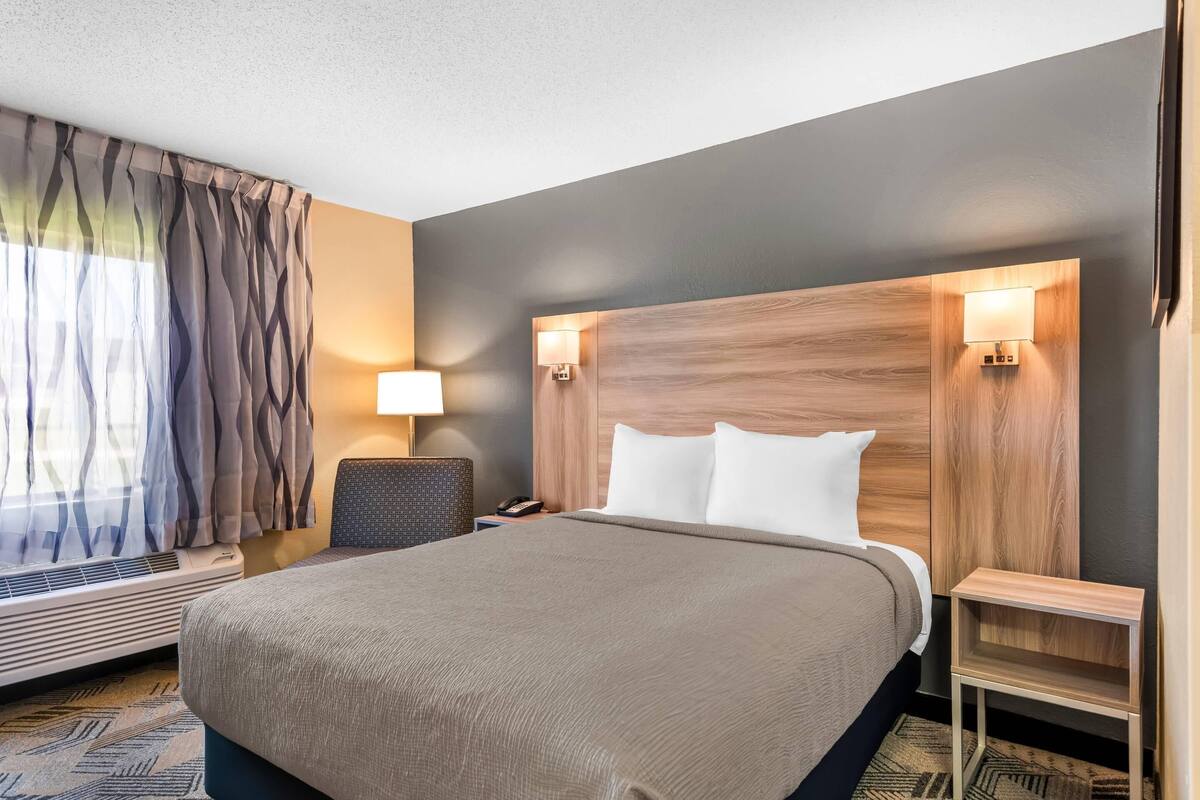 Een hotelkamer met een groot bed, twee bedlampen, een stoel, een nachtkastje en een raam met gordijnen.