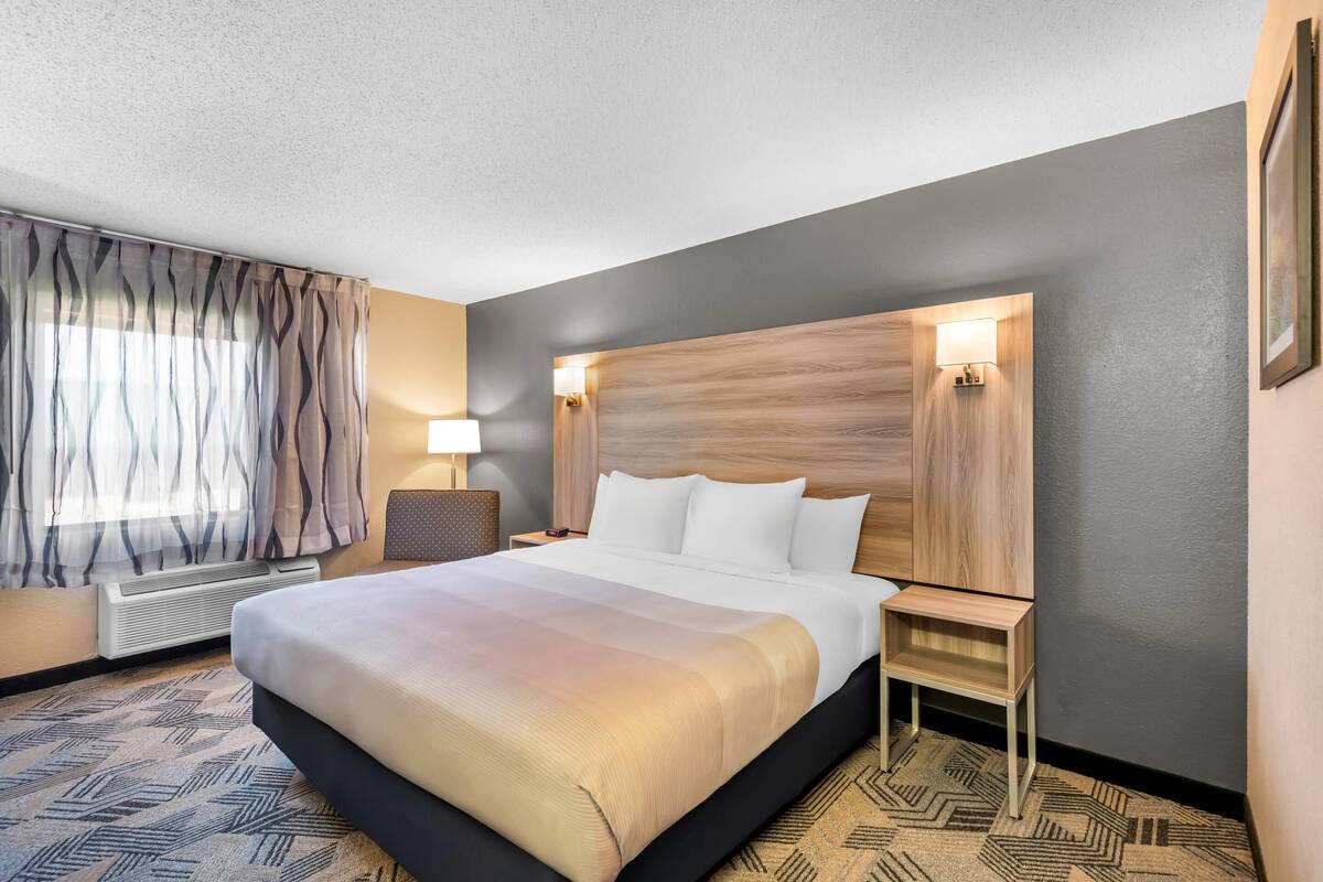Een hotelkamer met een groot bed, twee nachtkastjes, een wandlamp en een raam met gordijnen.