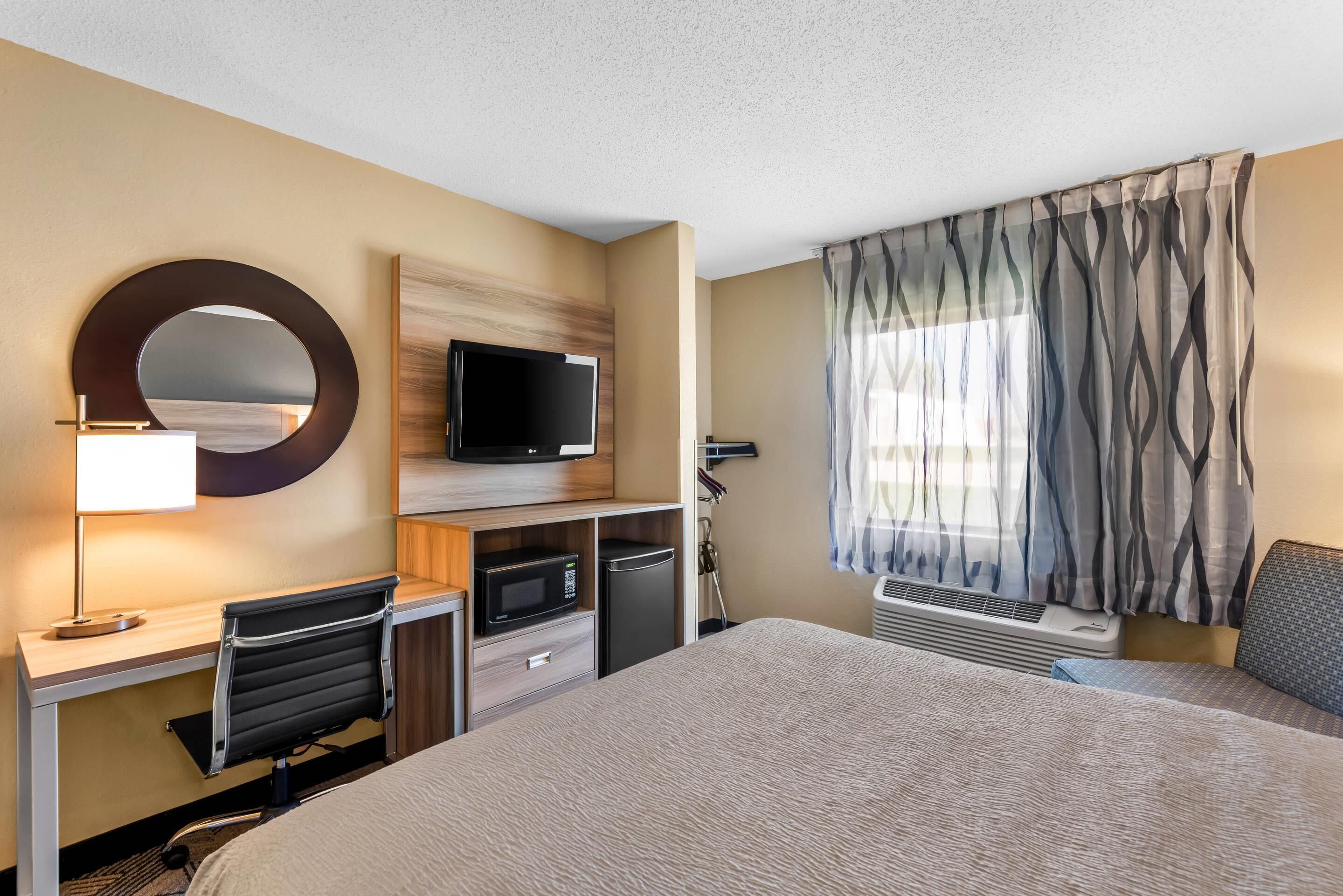 A hotel room with a bed, desk, chair, TV, microwave, and air conditioning unit.