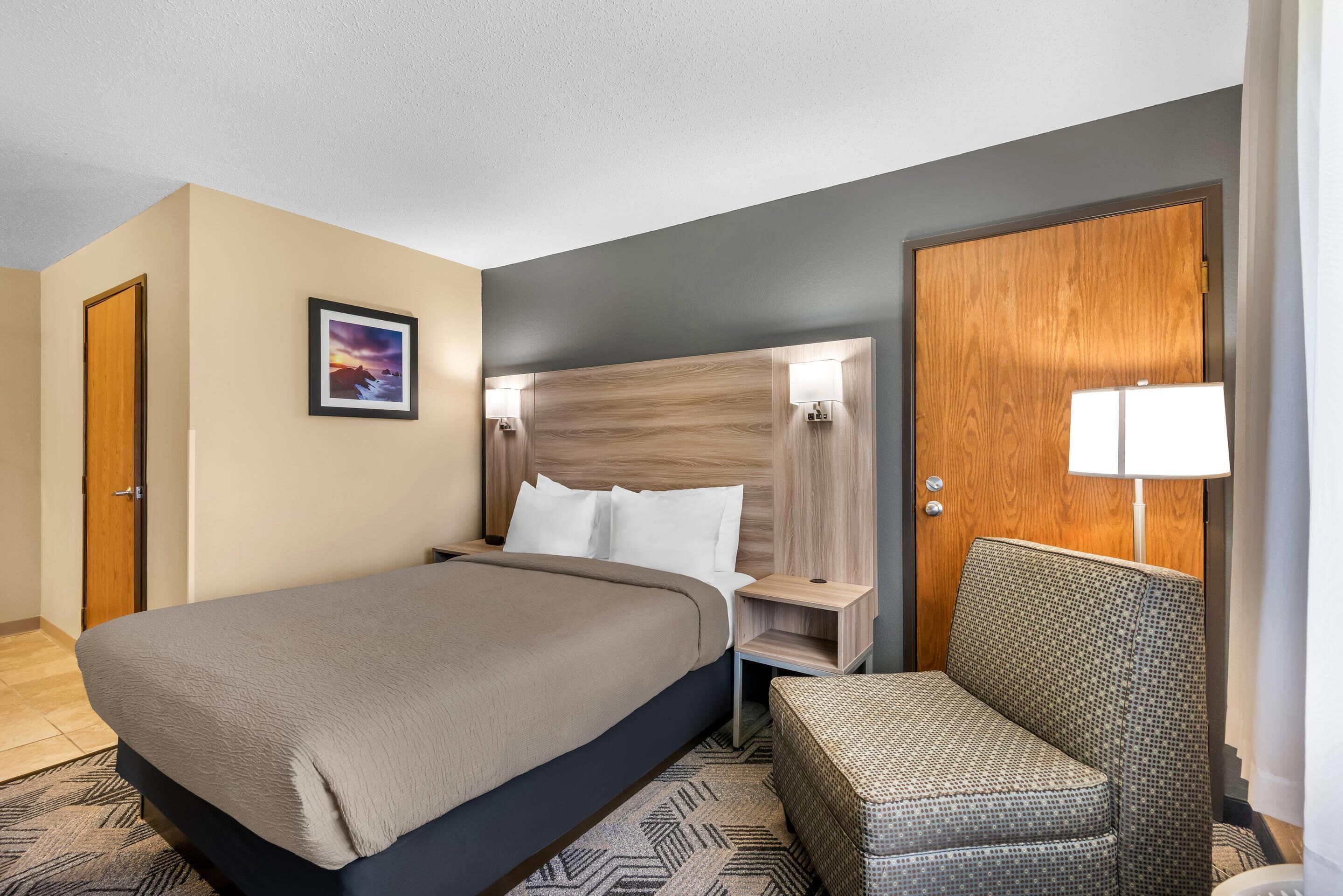 Habitación de hotel con cama, una silla, mesita de noche, cabecera de madera, un cuadro en la pared y una puerta.