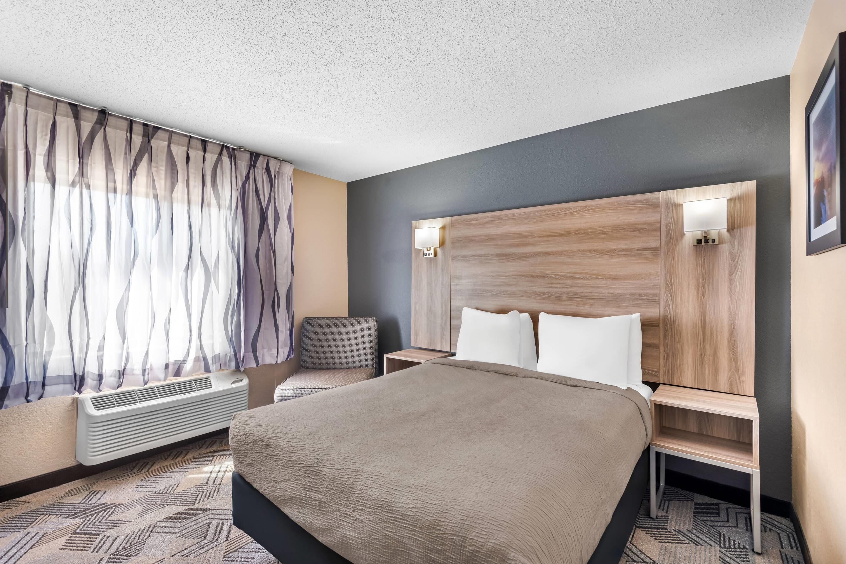 Habitación de hotel con una cama grande, una silla, una mesita de noche, una lámpara de pared y un ventanal con cortinas de gasa.