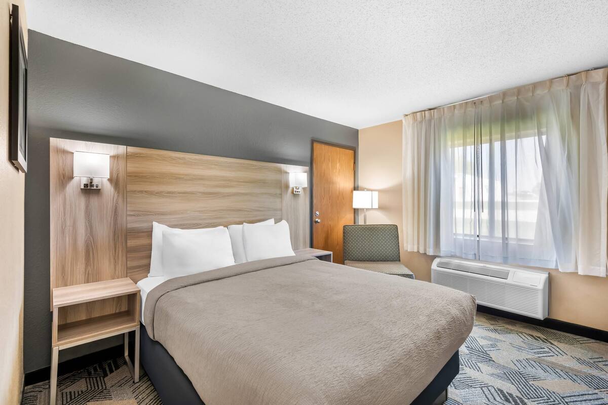 Een hotelkamer met een bed, nachtkastjes, een stoel, een lamp, een raam met gordijnen en een airconditioning.