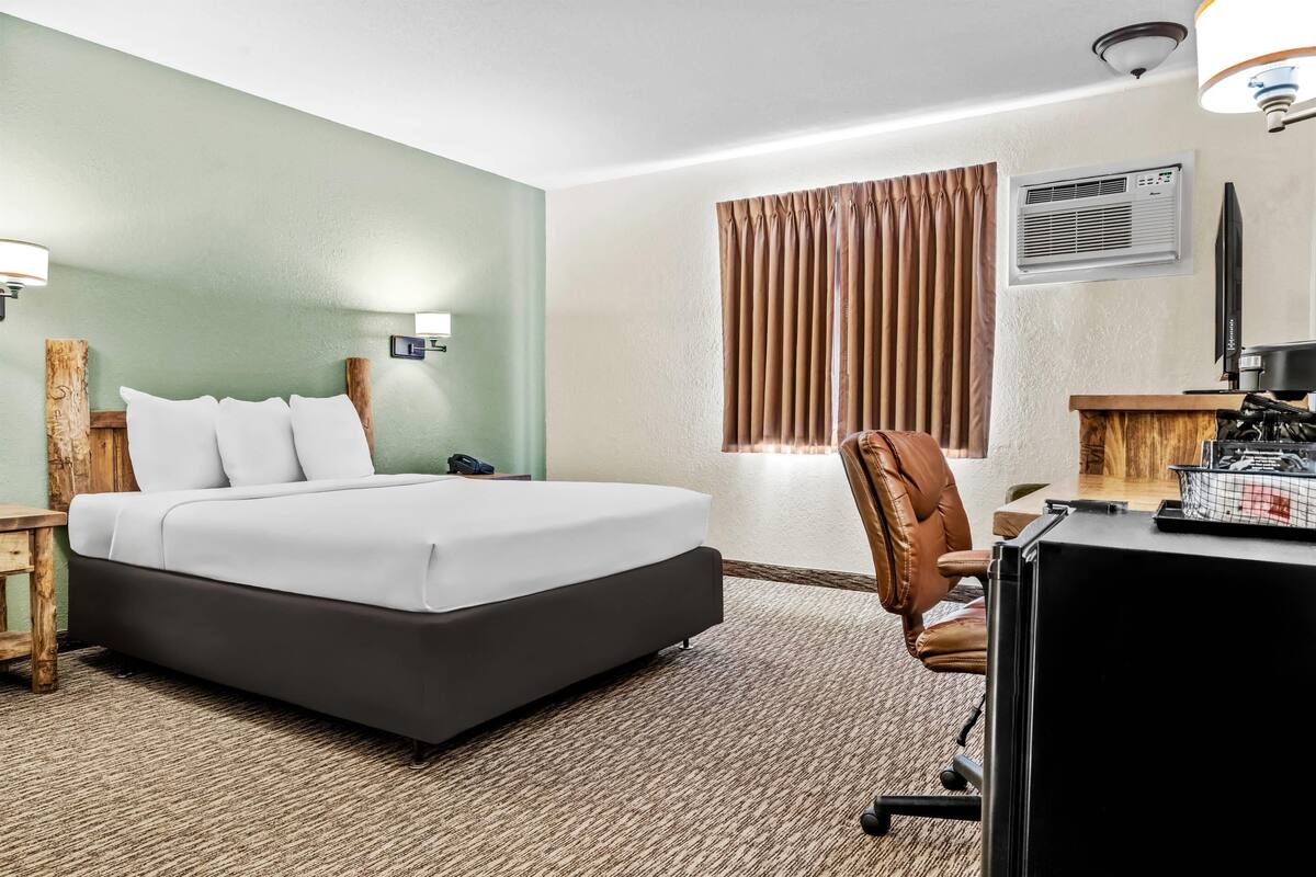 A hotel room with a bed, a desk, a chair, a lamp, an air conditioner, and a window with curtains.