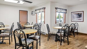 Uma sala de jantar com mesas e cadeiras redondas, um ventilador de teto e janelas com cortinas.