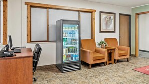 Uma área de recepção com uma mesa de madeira, um computador, uma geladeira abastecida com bebidas e duas cadeiras laranja.