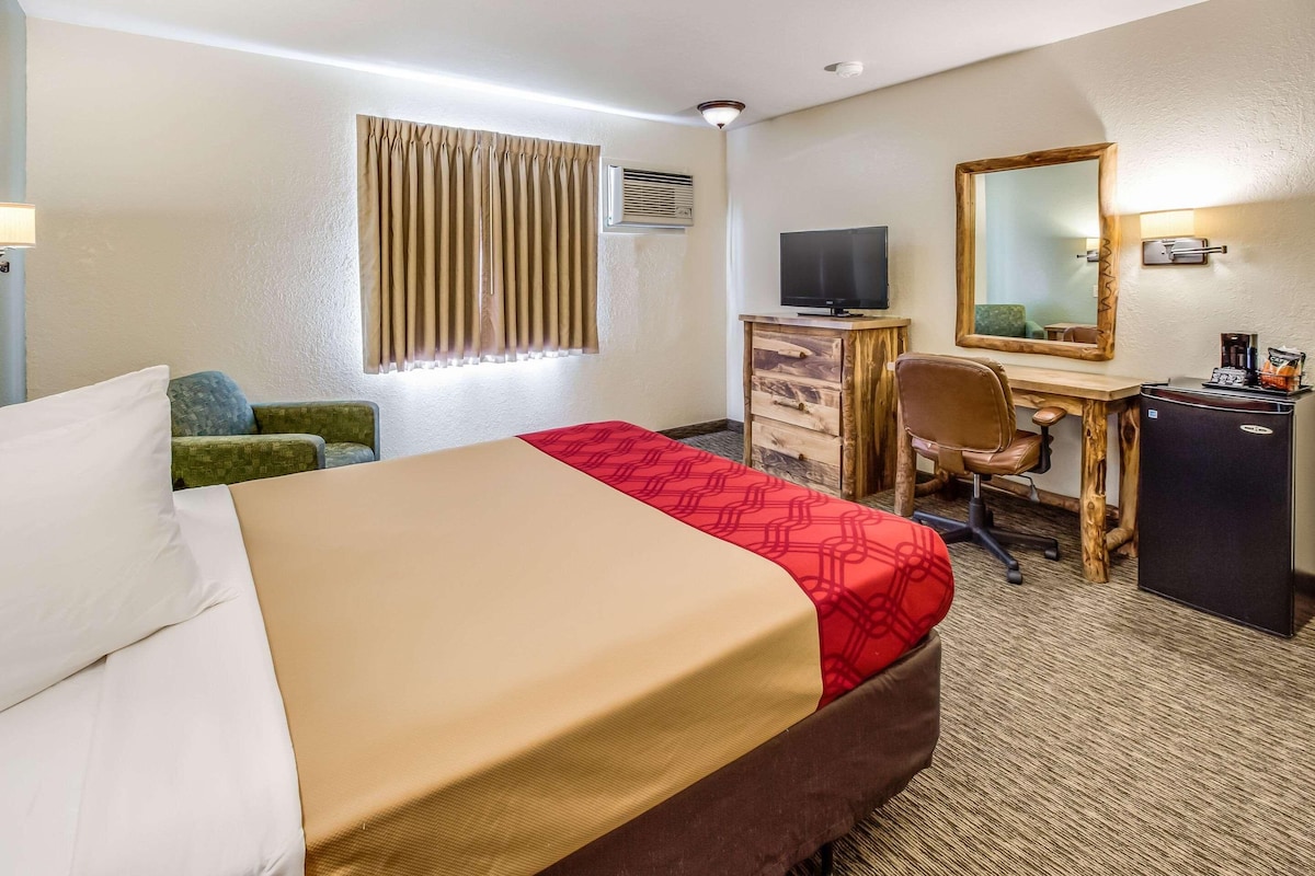 A hotel room with a bed, a desk with a computer, a chair, a dresser, a mirror, and a coffee machine.