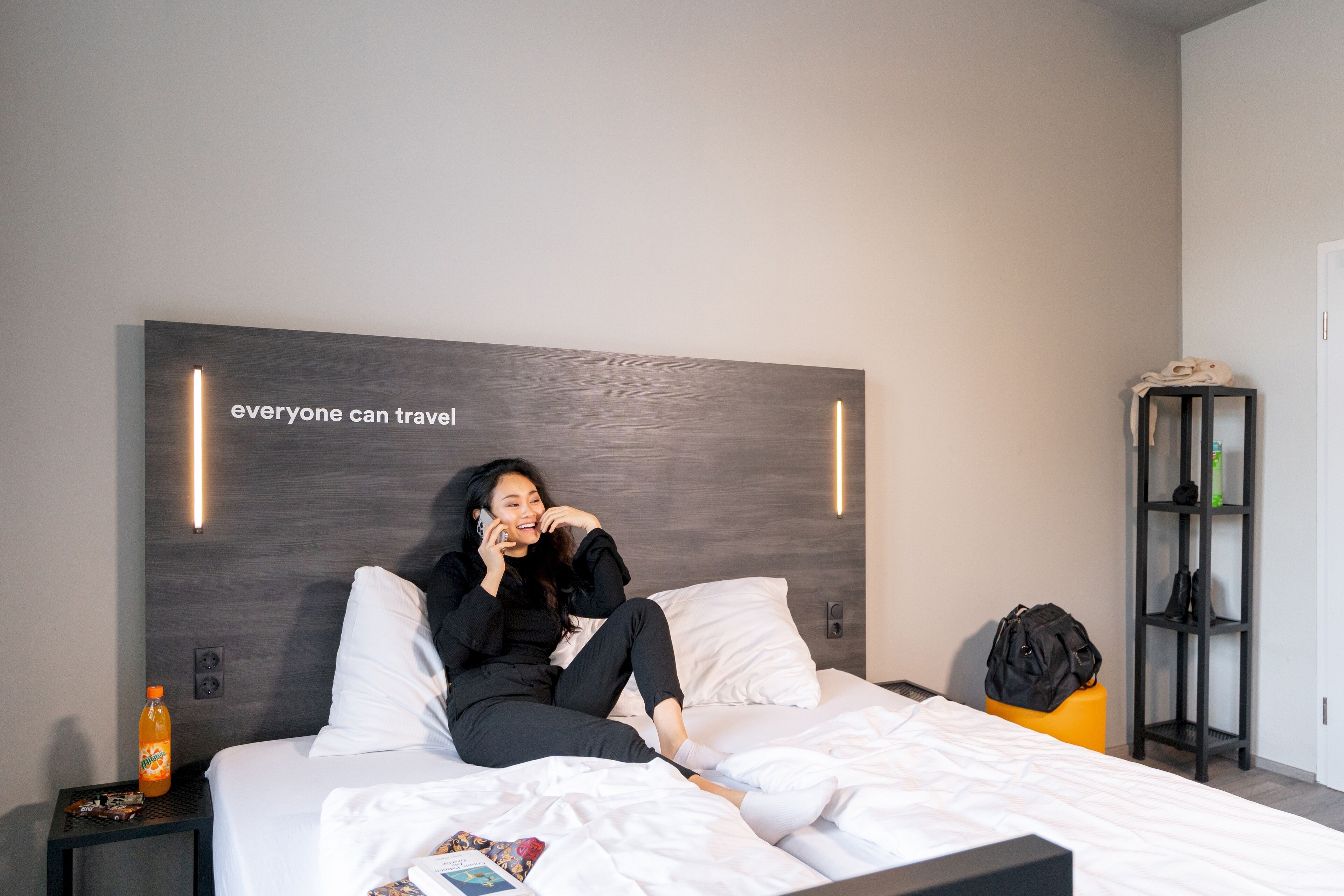A modern hotel room with a large bed, a nightstand with a bottle, and a bookshelf.