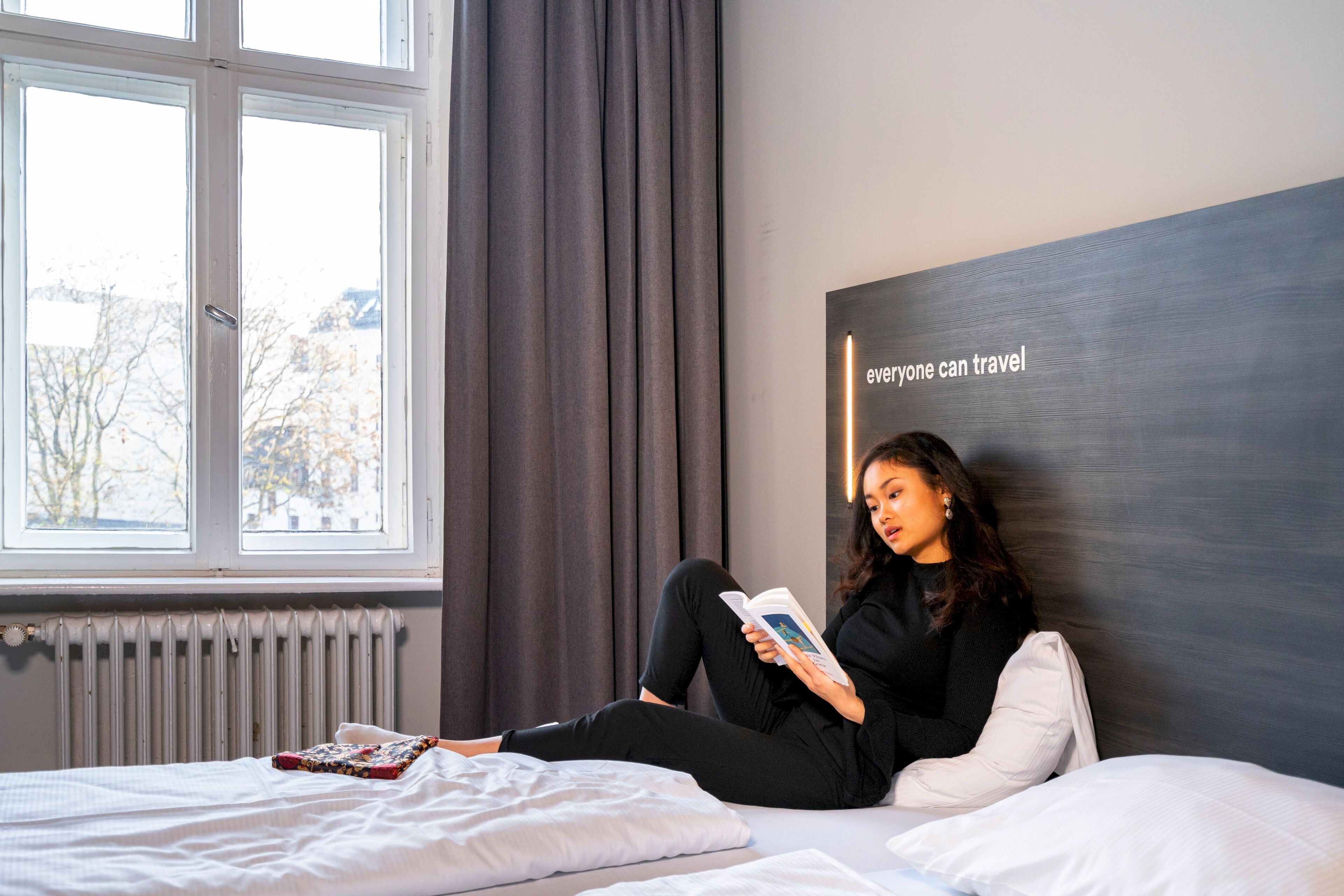 Una persona sentada en una cama leyendo un libro en una habitación con vistas a árboles a través de una ventana.