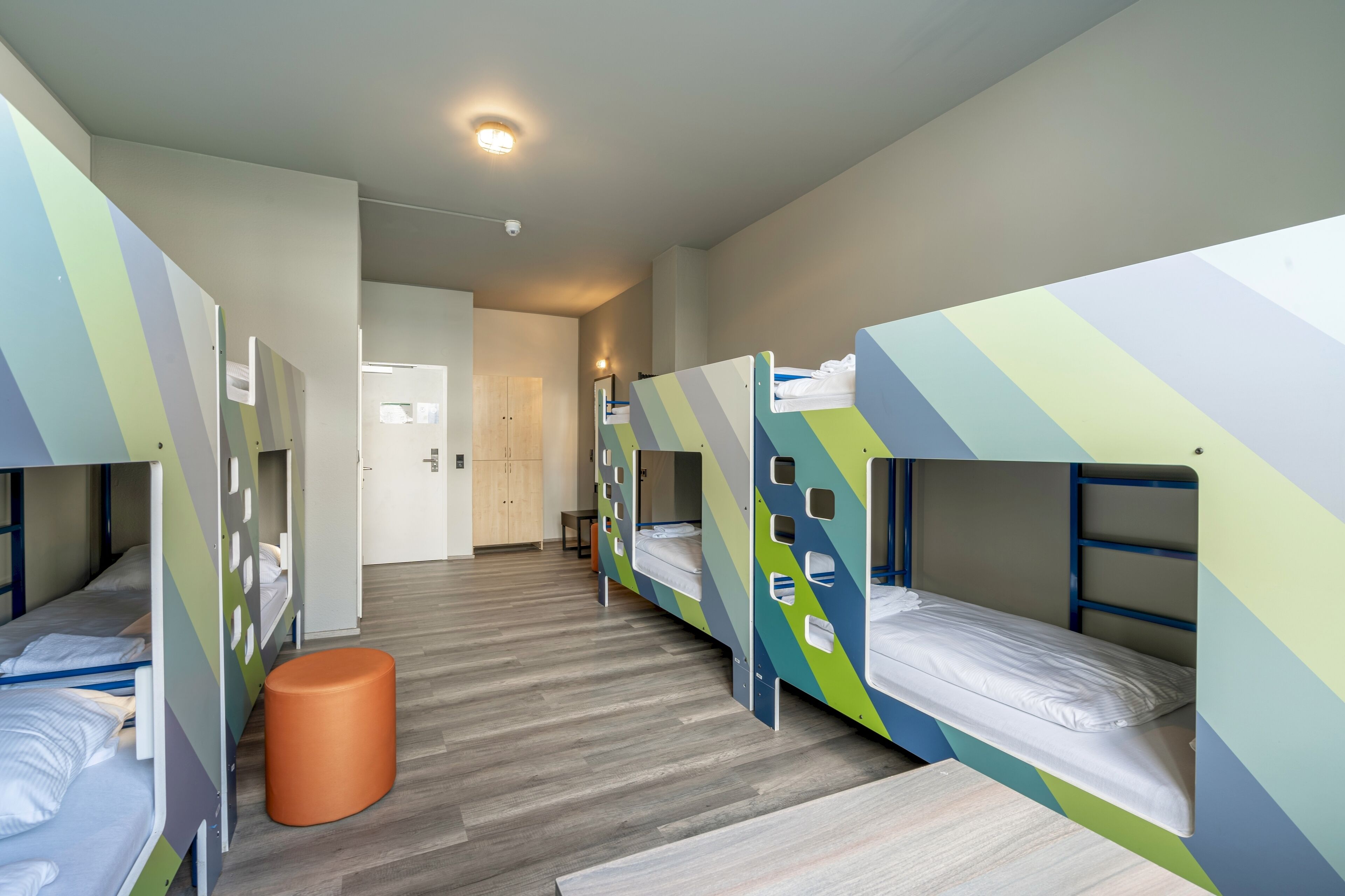 A dormitory room with bunk beds, a wooden floor, and a small orange stool.