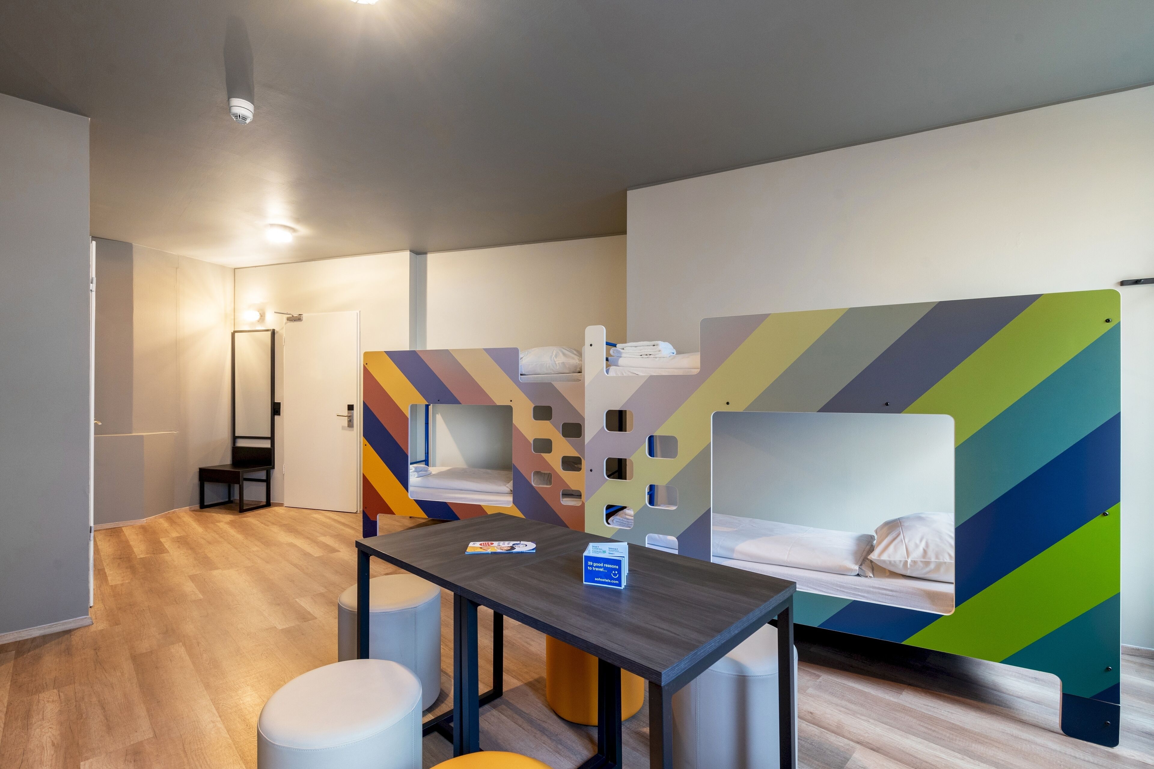 Ett modernt hotellrum med trägolv, ett bord i mitten med ett tidningsställ och en låda, och två våningssängar med väggar dekorerade med färgglada, geometriska mönster.