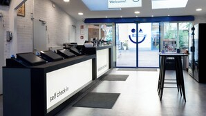 A self-check-in counter with a digital display, a reception area with a glass door, and a view of the outside. - a&o Berlin Friedrichshain (Berlin)