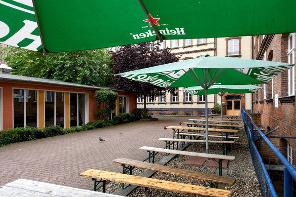 An outdoor patio area with picnic tables and green umbrellas, surrounded by brick buildings and greenery. - a&o Berlin Friedrichshain (Berlin)