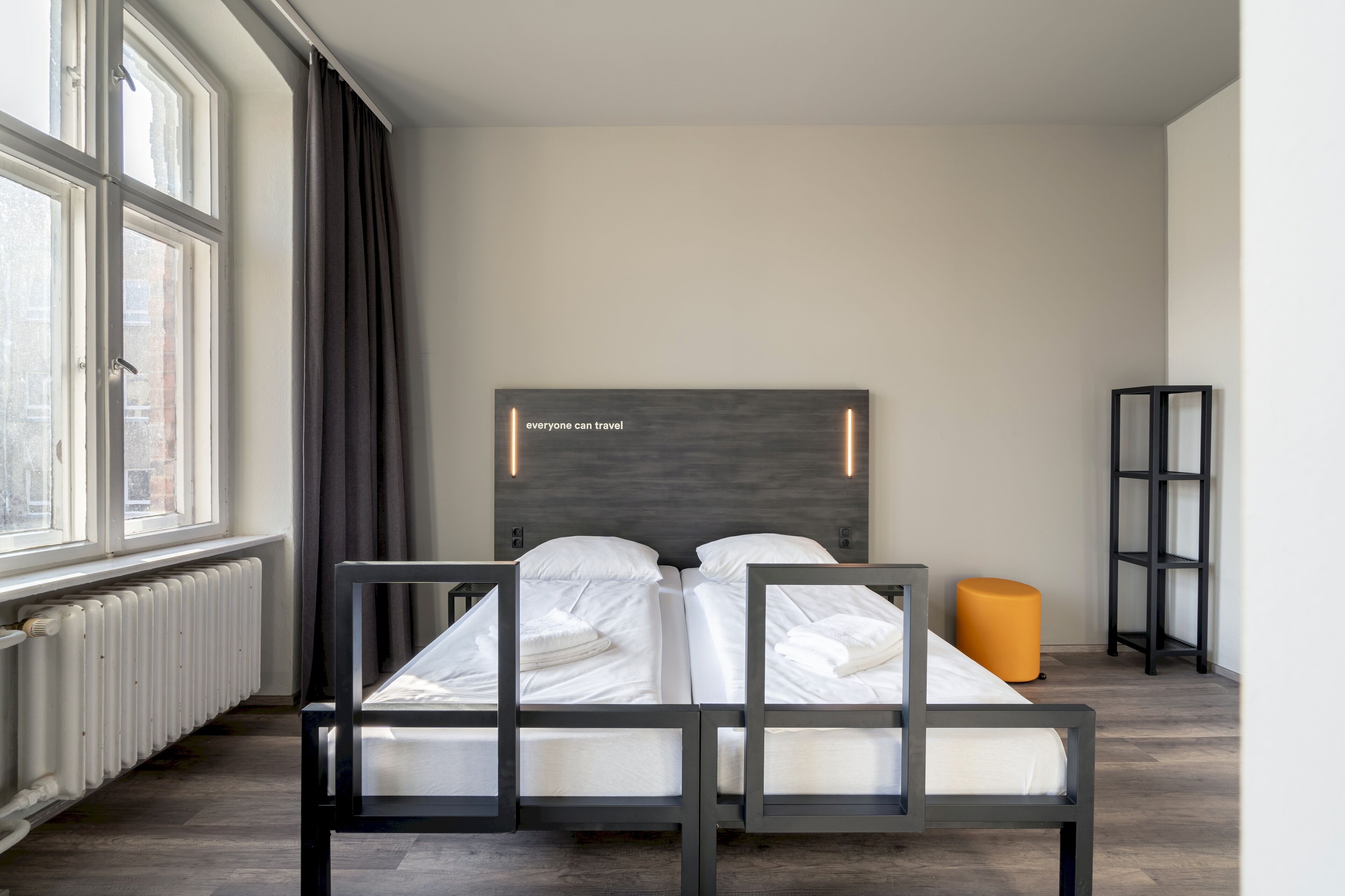 Ein modernes Hotelzimmer mit einem Doppelbett, einer Kopfstütze mit einem motivierenden Zitat, einem Heizkörper, einem Fenster mit Vorhängen und einem kleinen orangefarbenen Hocker.