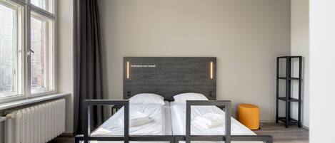 Una habitación de hotel moderna con una cama doble, un cabecero con una cita motivacional, un radiador, una ventana con cortinas y un taburete naranja pequeño.