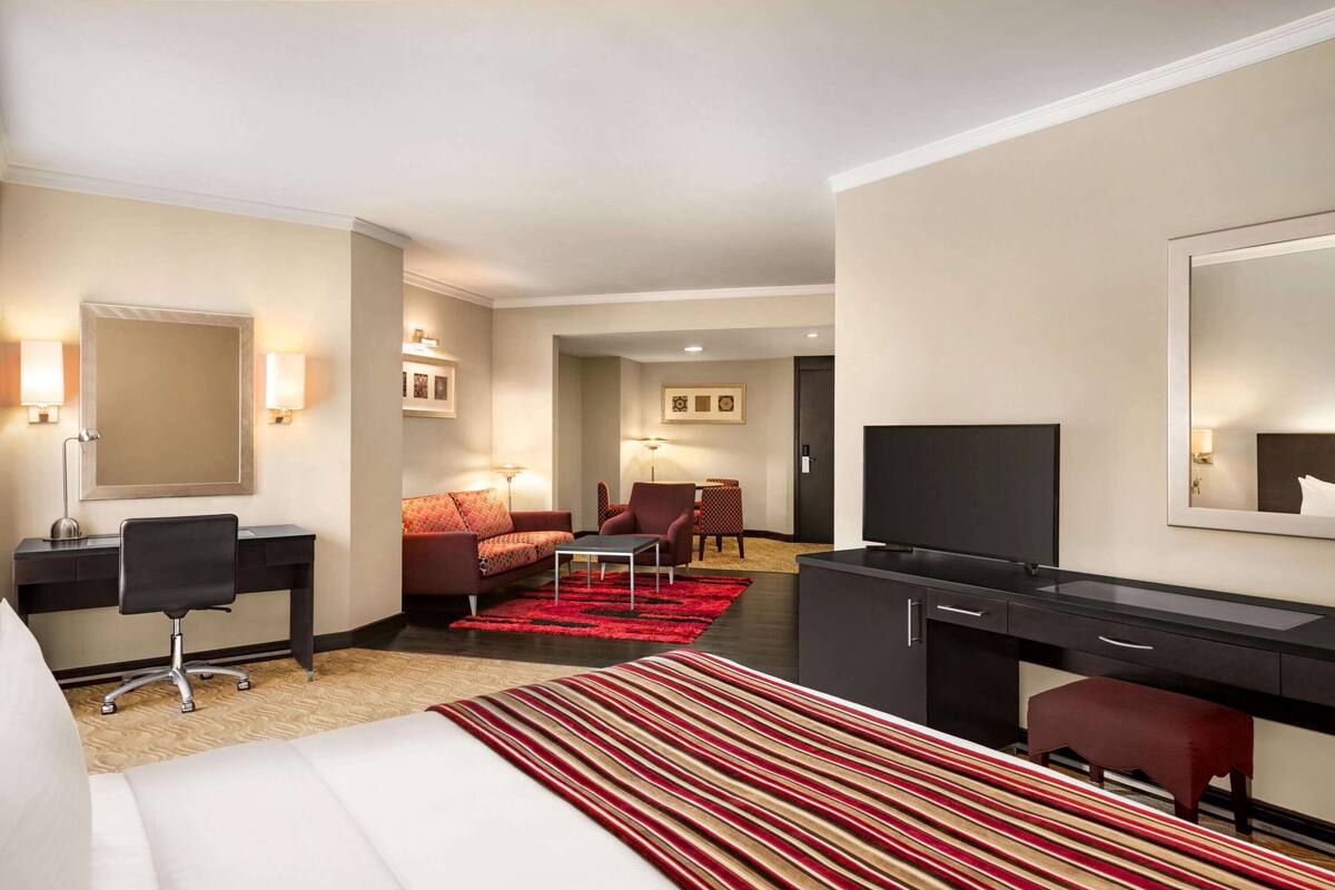 A hotel room with a bed, desk, chair, TV, and a red and white striped area rug.