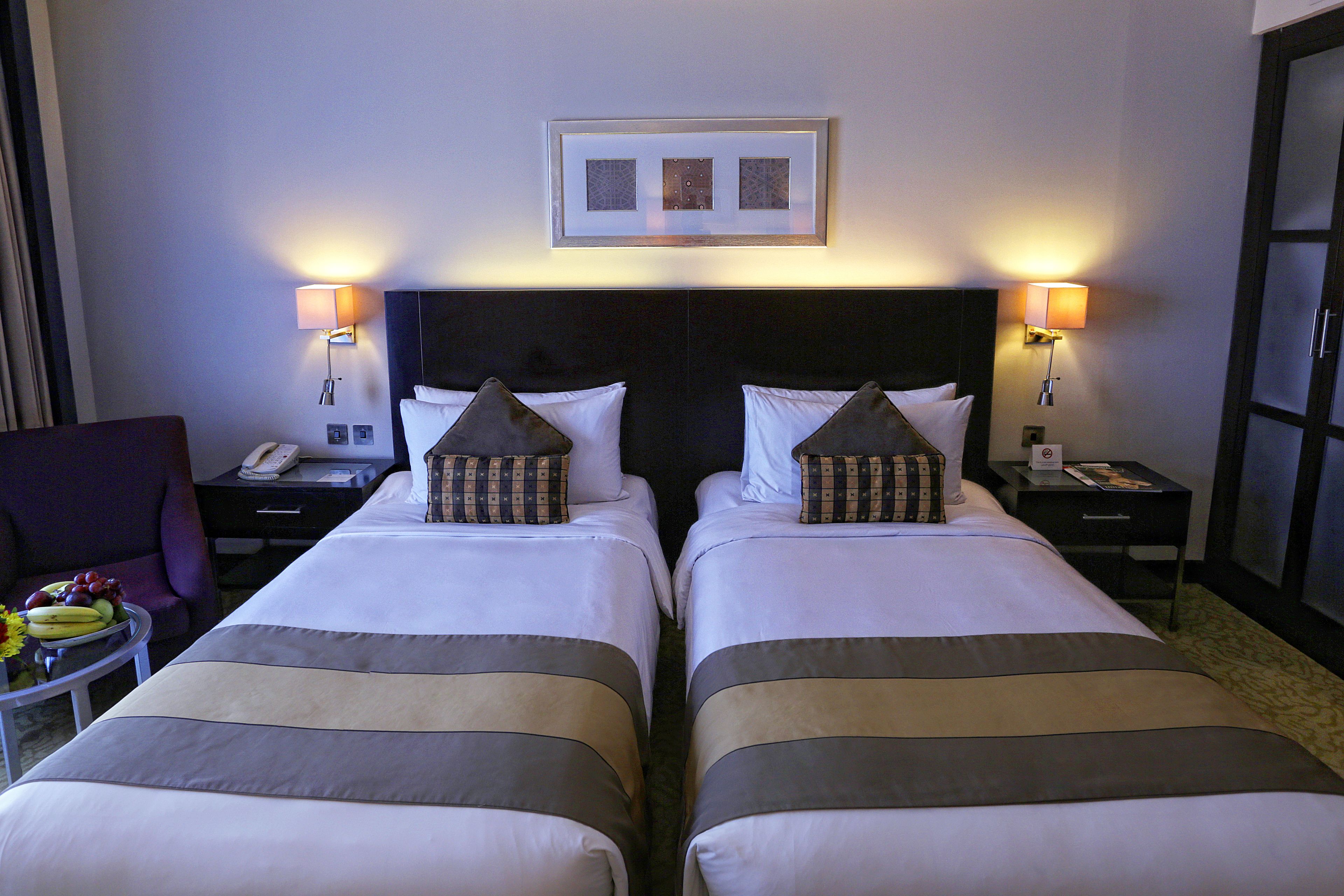Een hotelkamer met twee bedden, een nachtkastje met een telefoon, een stoel en een ingelijste foto aan de muur.
