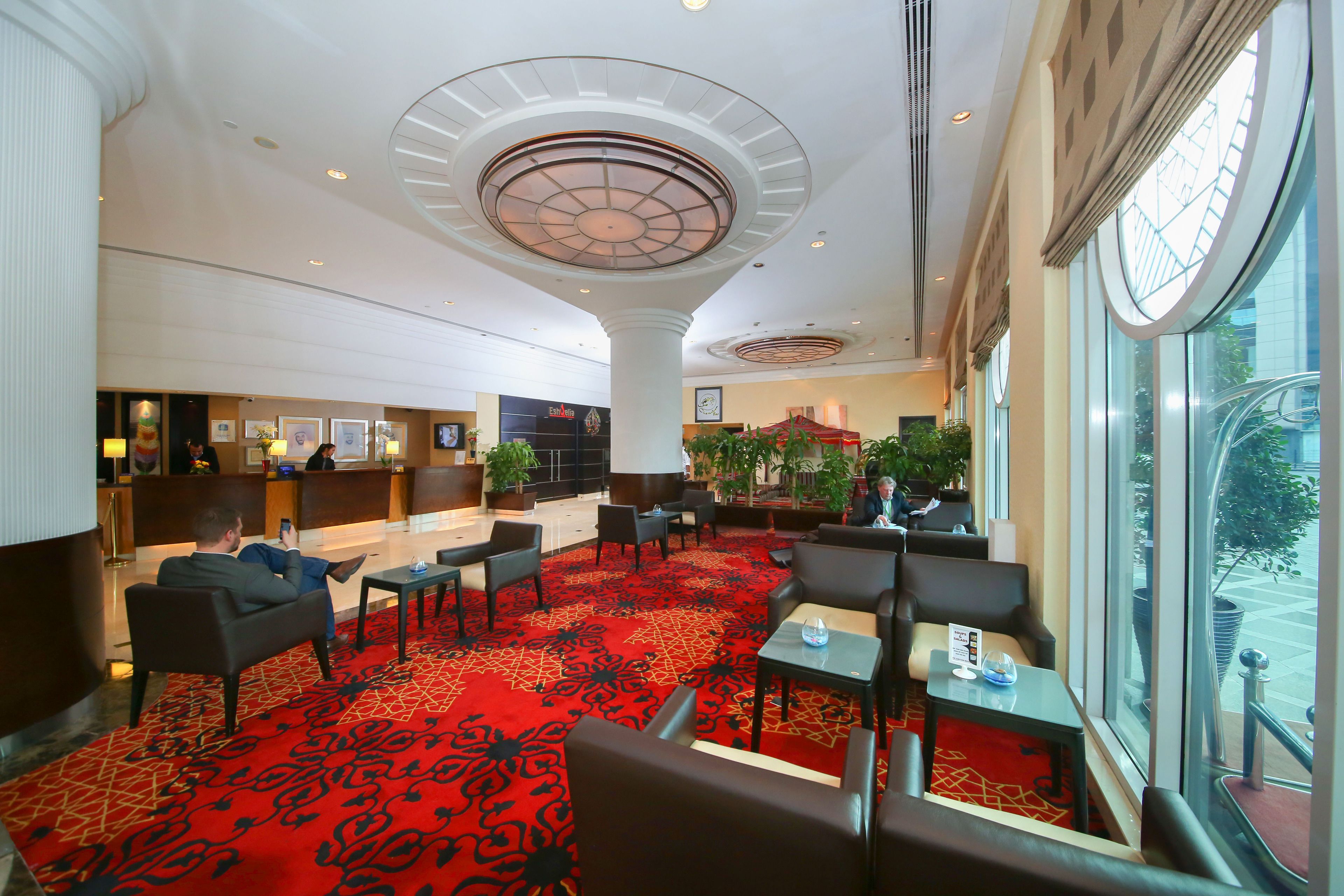 A hotel lobby with a red carpet, seating areas, a reception desk, and large windows offering a view outside.