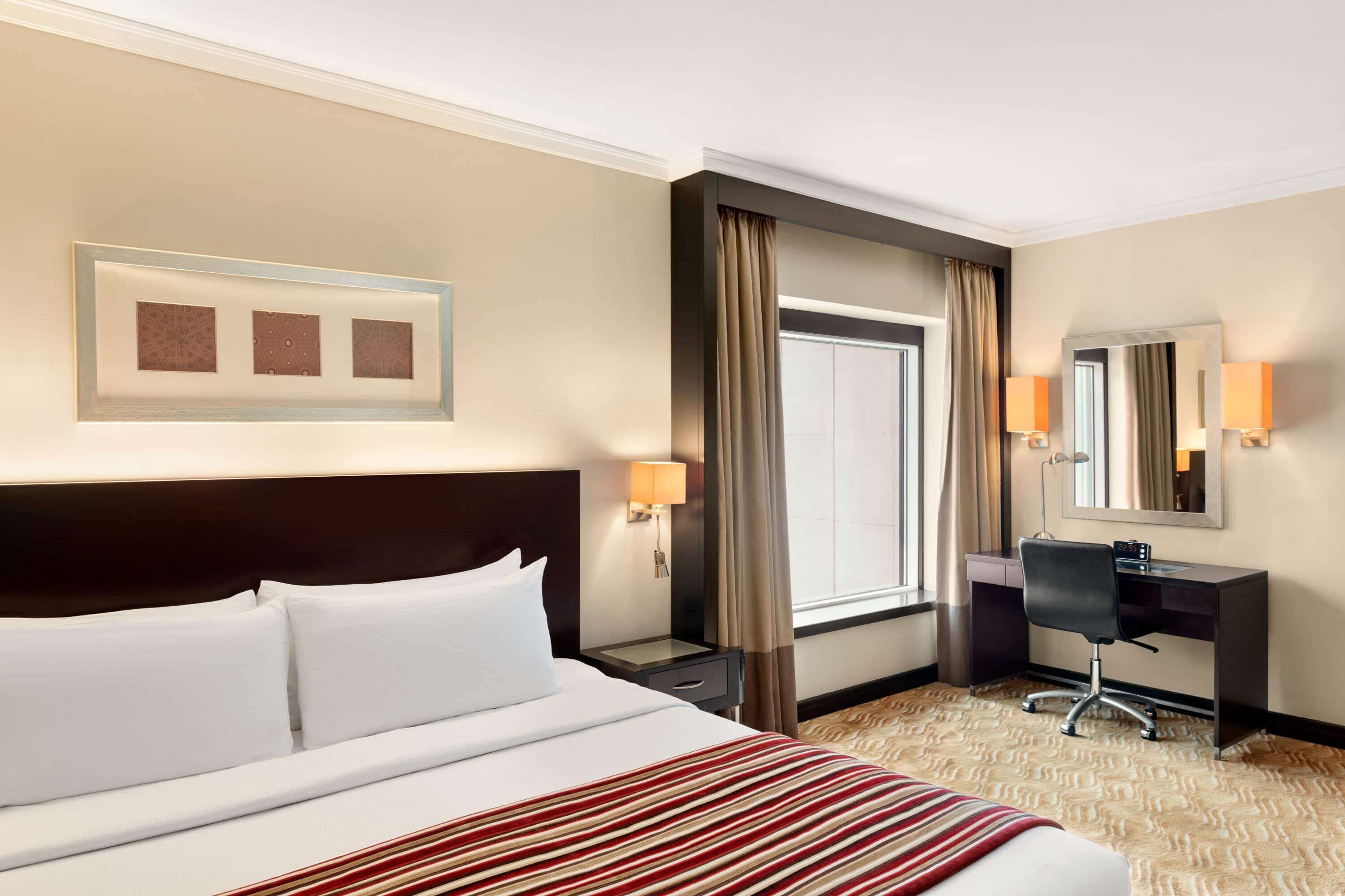 Ein Hotelzimmer mit einem großen Bett, einem Schreibtisch, einem Stuhl, einer Schreibtischlampe, einem Nachttisch, einem Spiegel und einem Fenster mit Vorhängen.