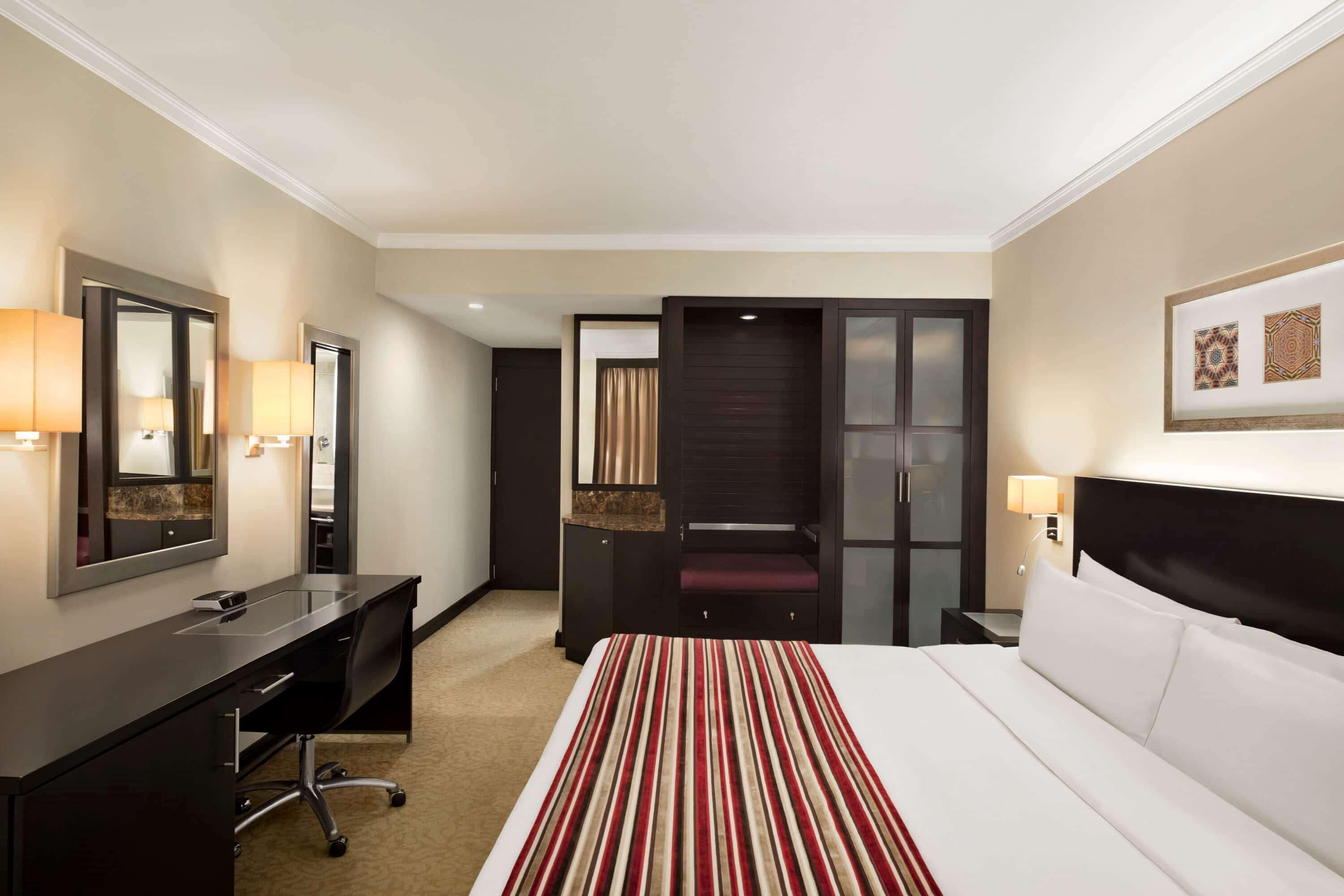 A hotel room with a bed, a desk, a chair, a wardrobe, and a mirror.