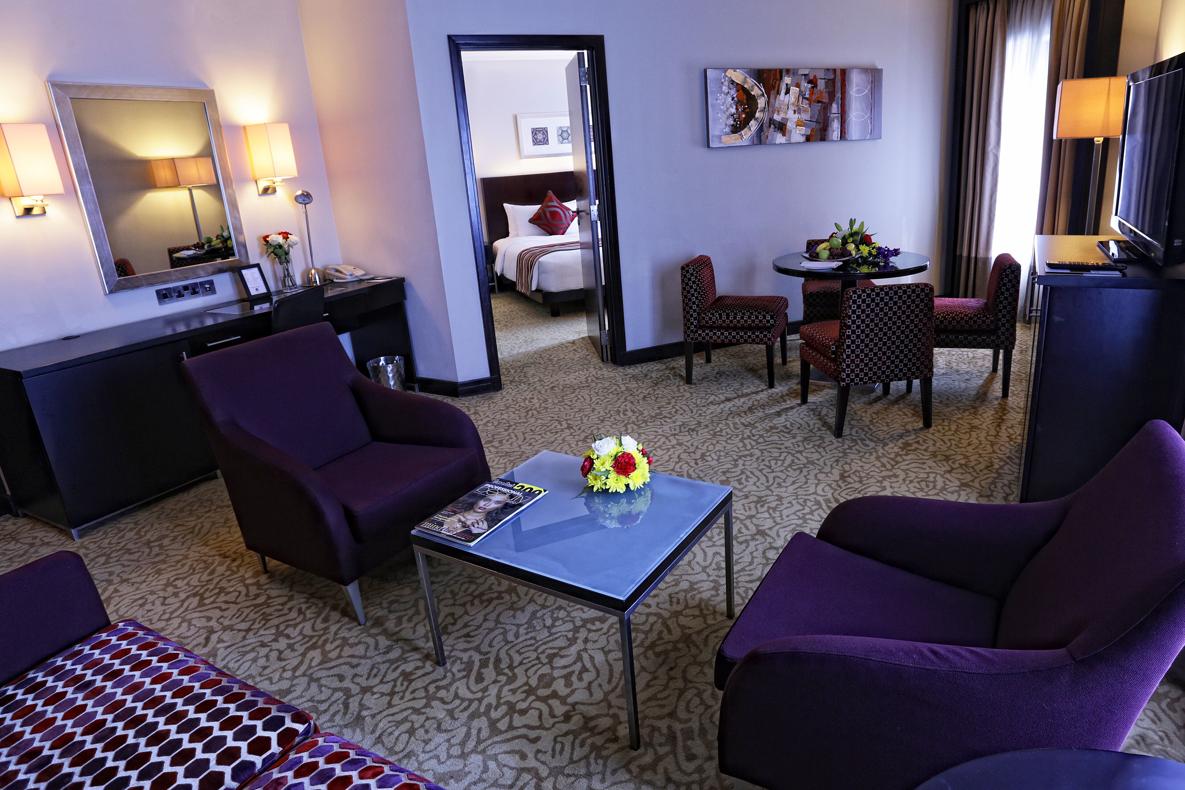 A hotel room with a bed, a desk, two purple armchairs, a small table with a vase of flowers, a television, and a large mirror.