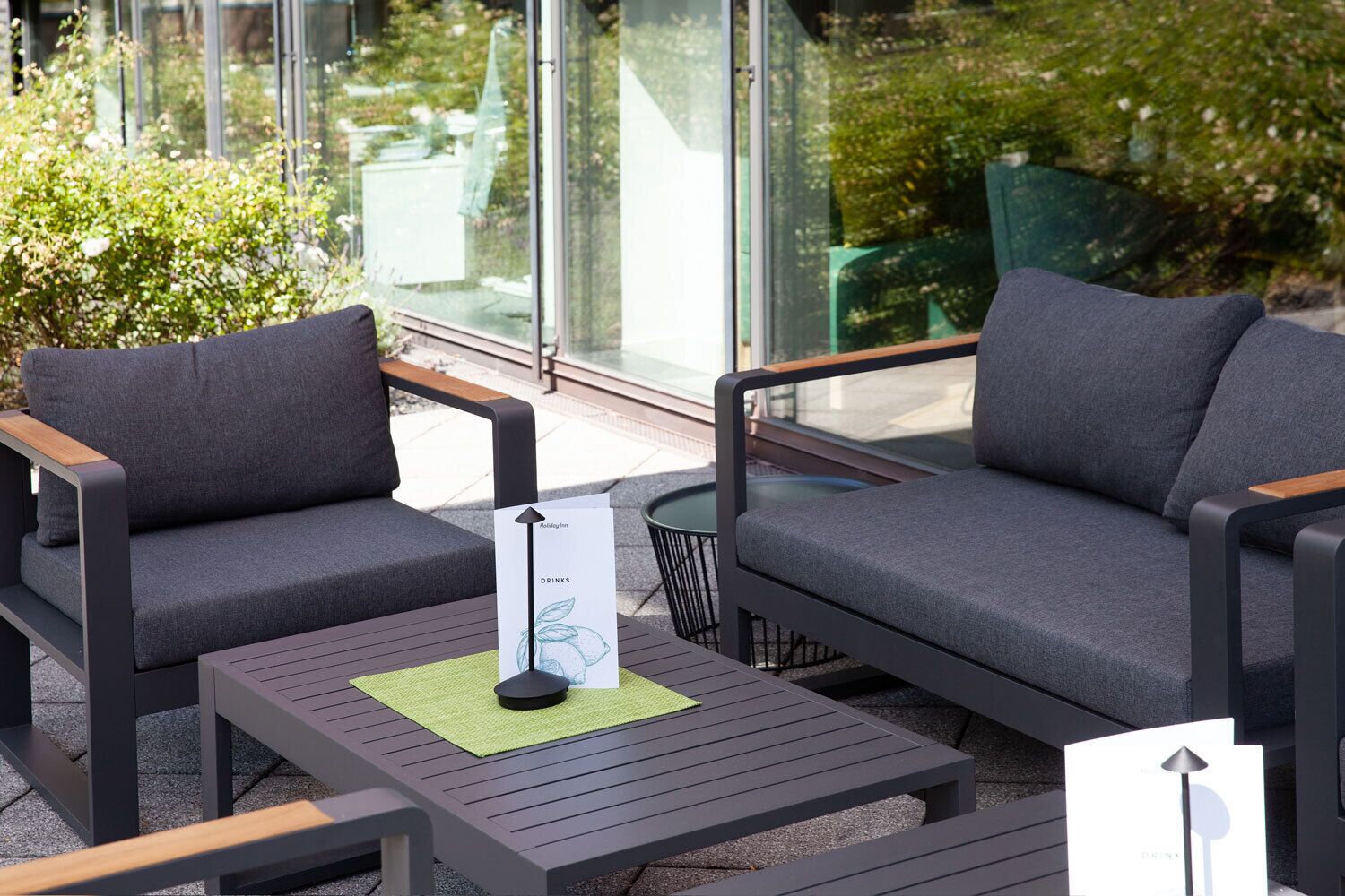 An outdoor seating area with two grey sofas and a small table, a menu sign, and a glass wall with greenery.