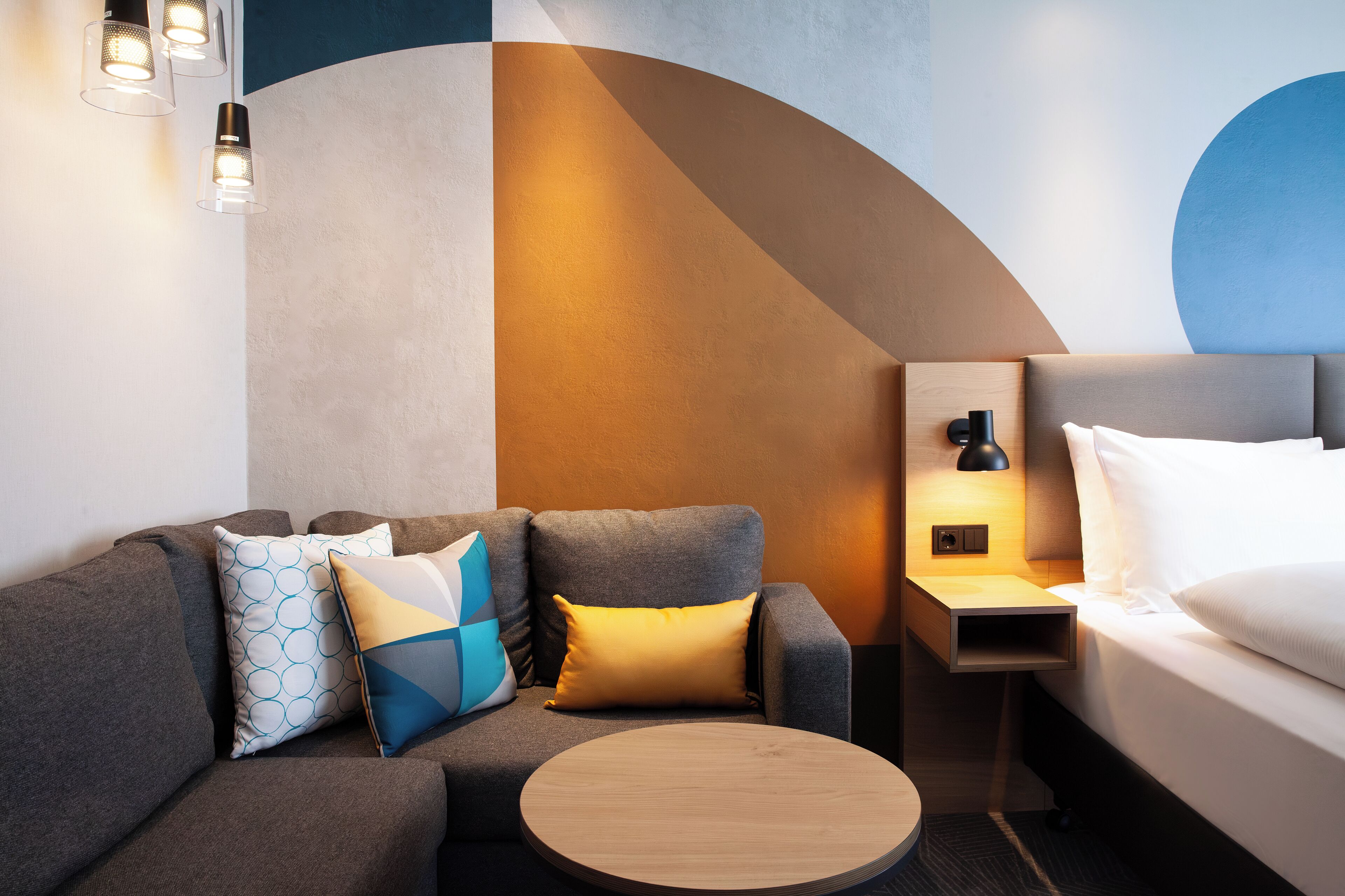 A modern hotel room with a sofa, a round wooden table, a bed, and a nightstand with a lamp.