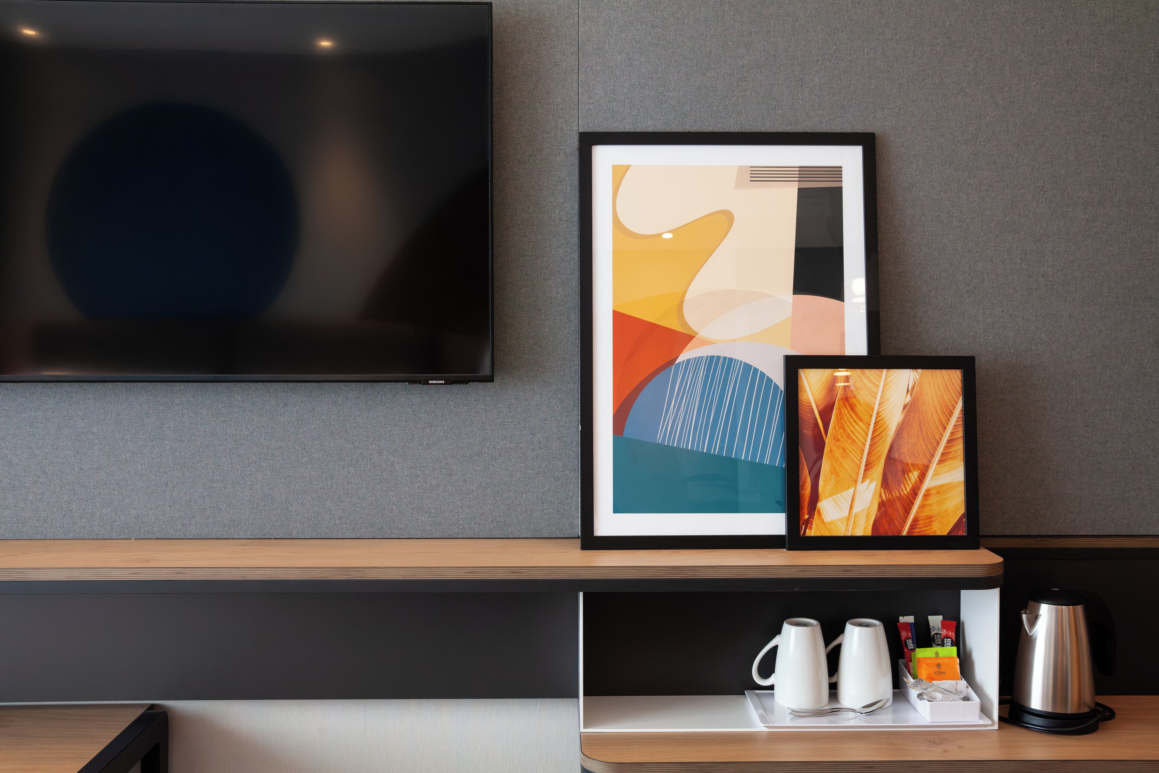 A modern hotel room with a flat-screen TV, abstract artwork, a coffee maker, and a tray with a mug and glasses.