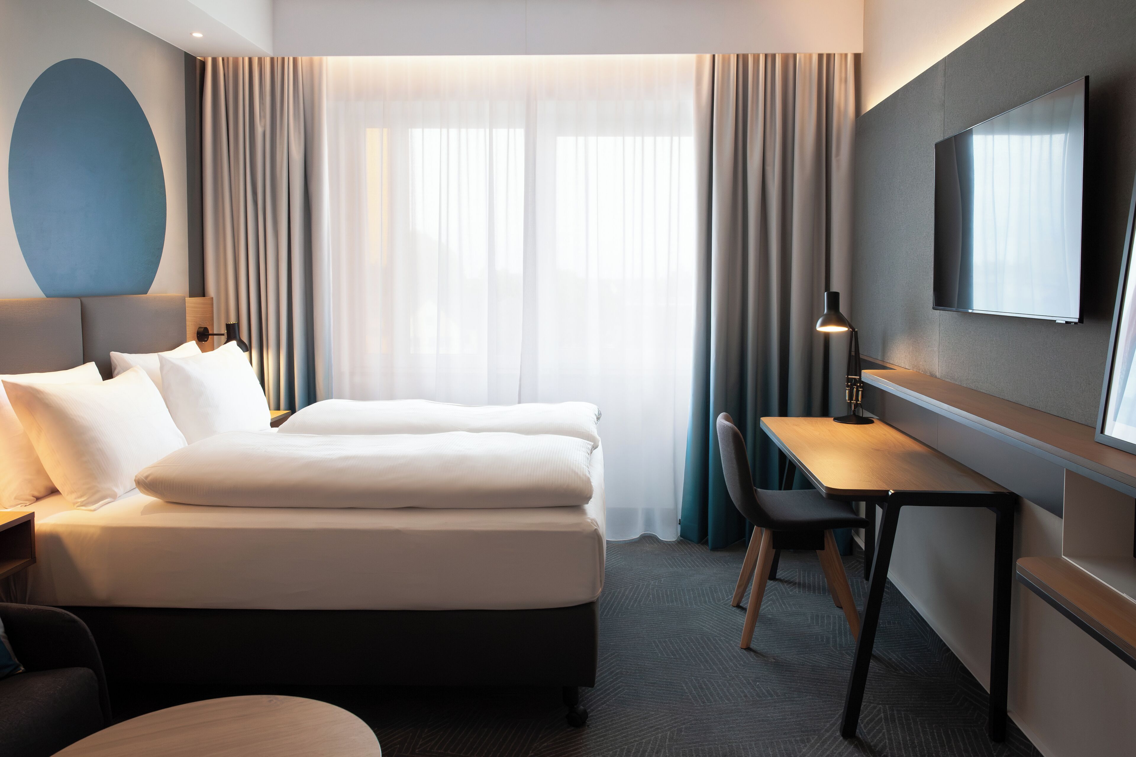 A modern hotel room with a bed, desk, chair, TV, and large windows with curtains.