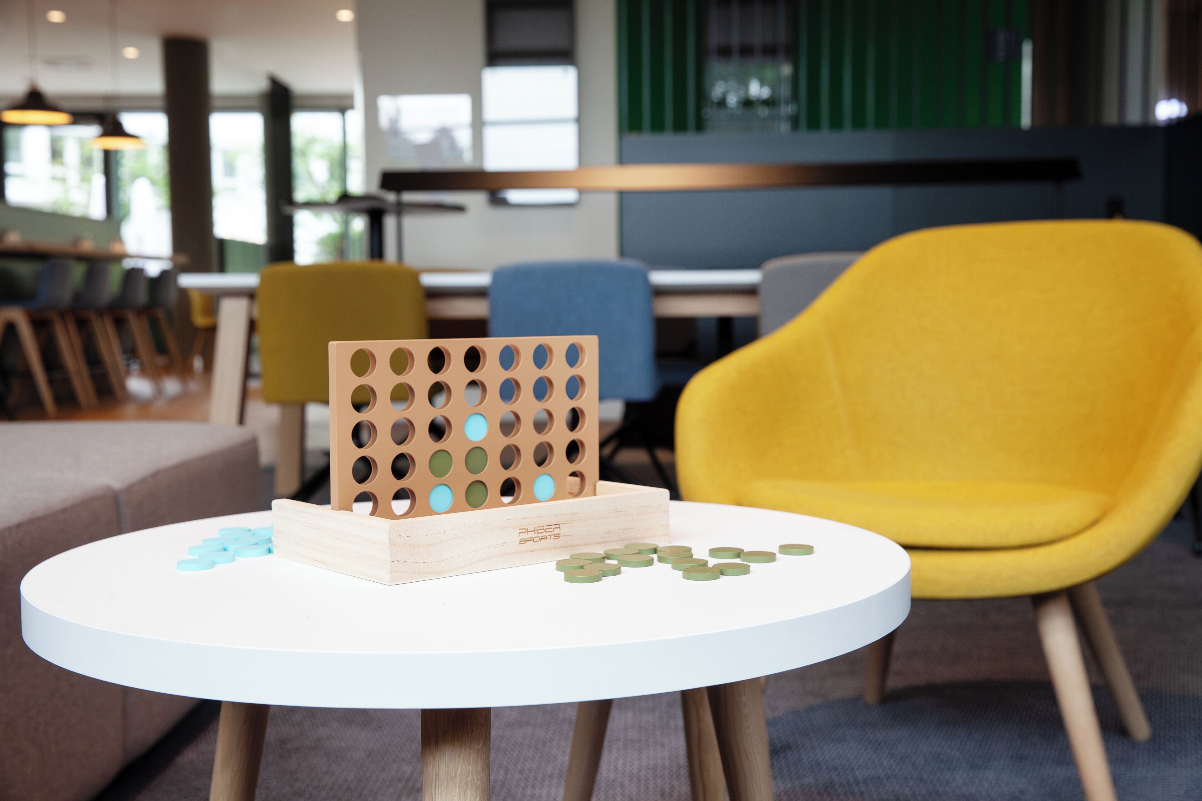 A round white table with a wooden pegboard and wooden blocks, a yellow chair, and a grey couch in a modern interior space.