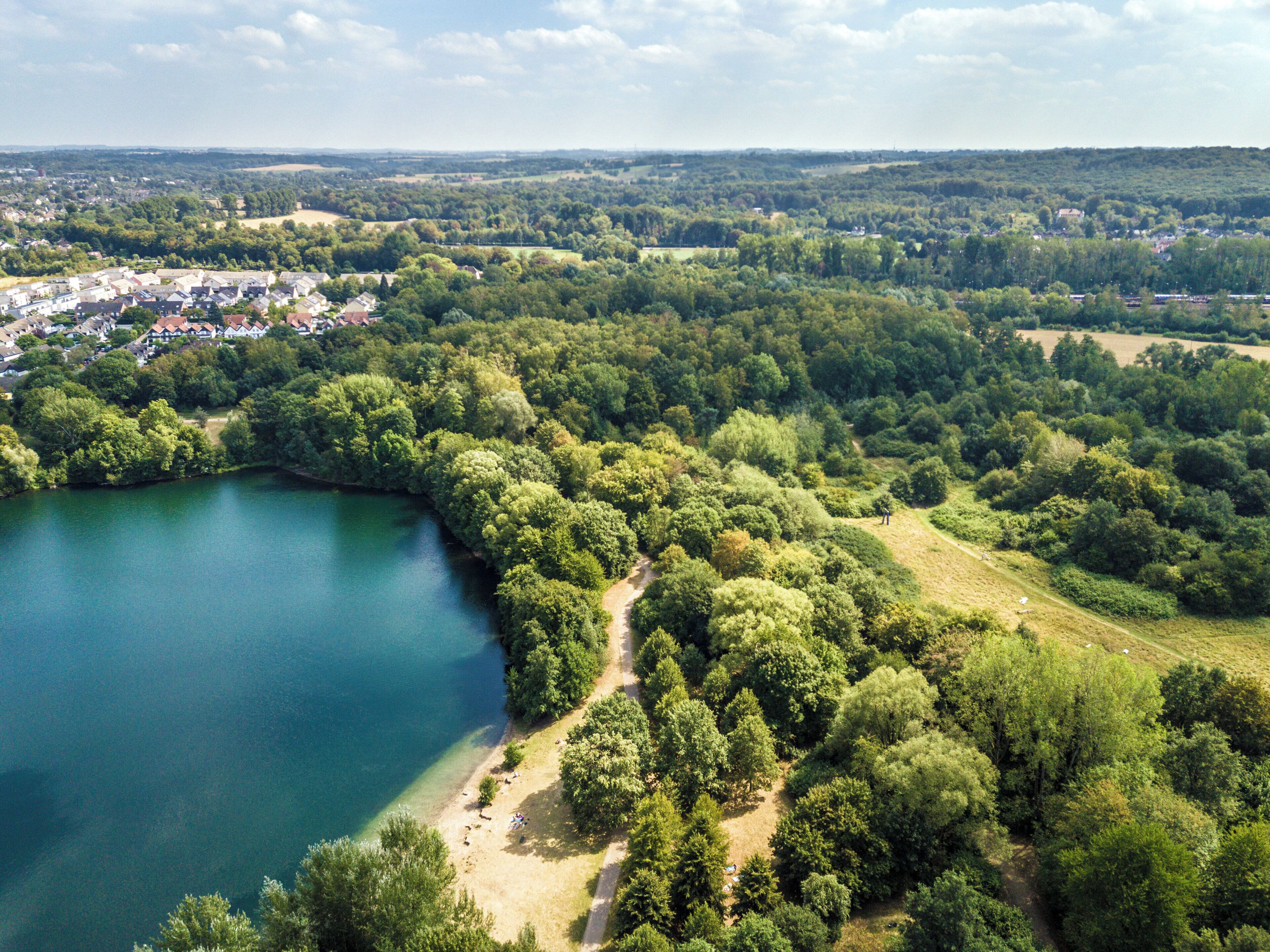 An aerial view of a lakeside area with dense greenery, a clear lake, and a residential area in the background.